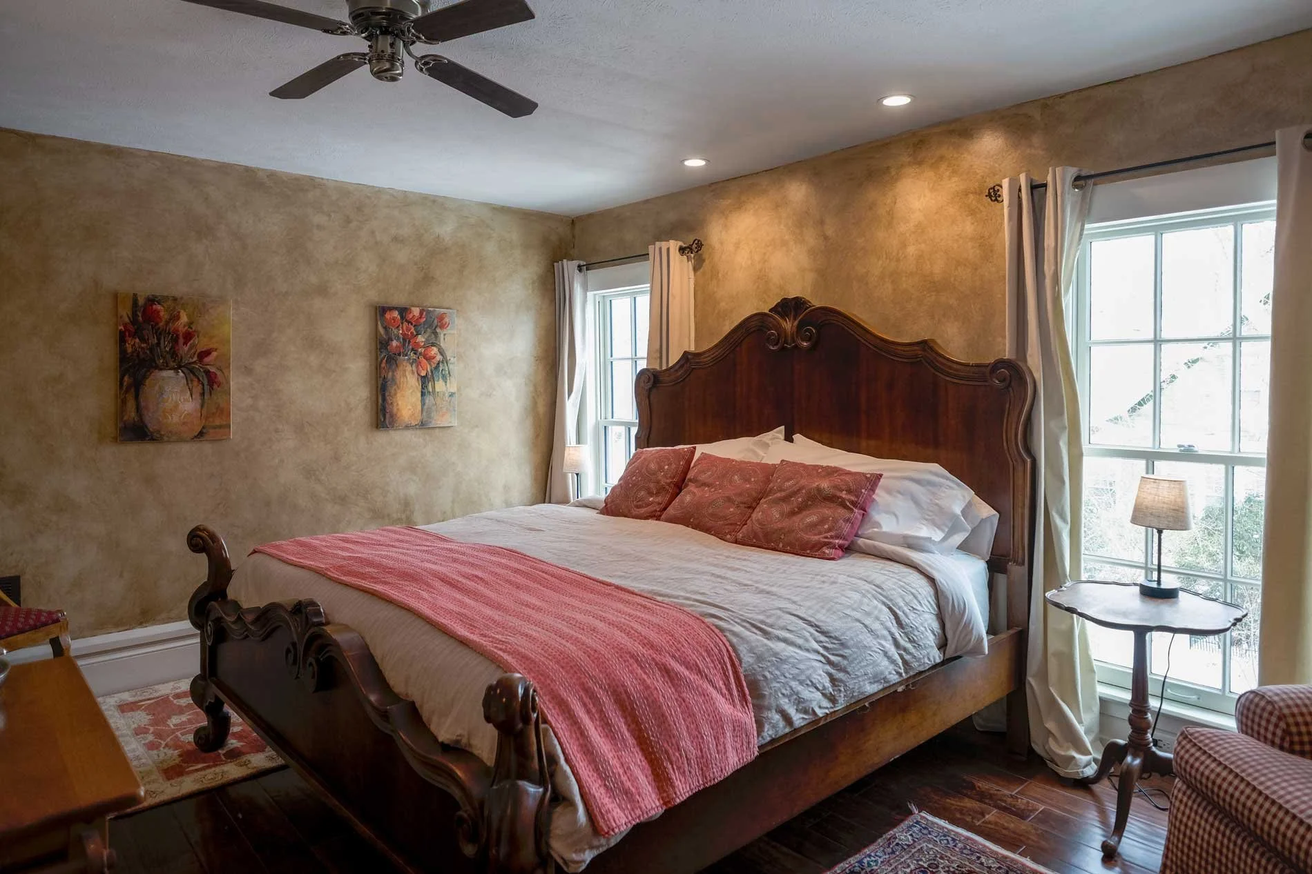 A bedroom with beige textured walls, a large wooden bed with a high ornate headboard, white bedding, and pink pillows. Two windows with white curtains, a small round side table with a lamp, and a ceiling fan.