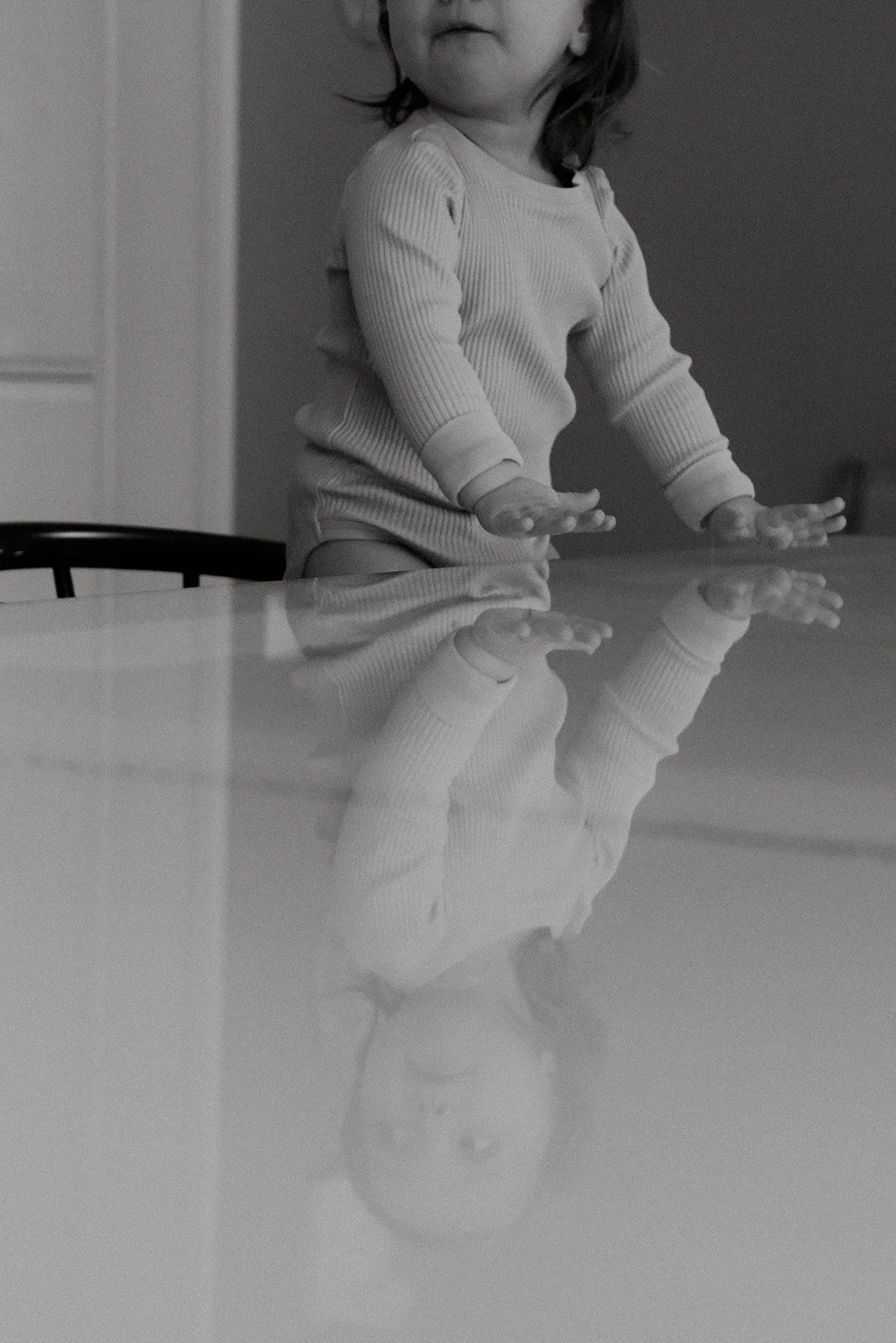 Mustavalkoisessa kuvassa pöydän pintaan heijastuu lapsen kuva. Lapsikuvaus kotona, kuvaaja Heidi Väisänen.