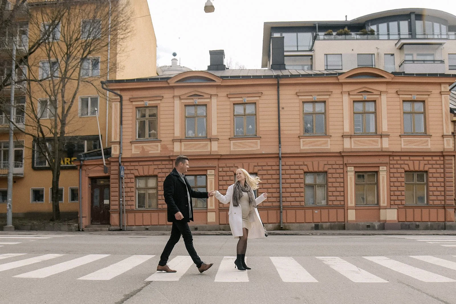 Nauravainen pariskunta ylittää tietä kaupunkimaisemassa. Pariskuntakuvaus Kerava.