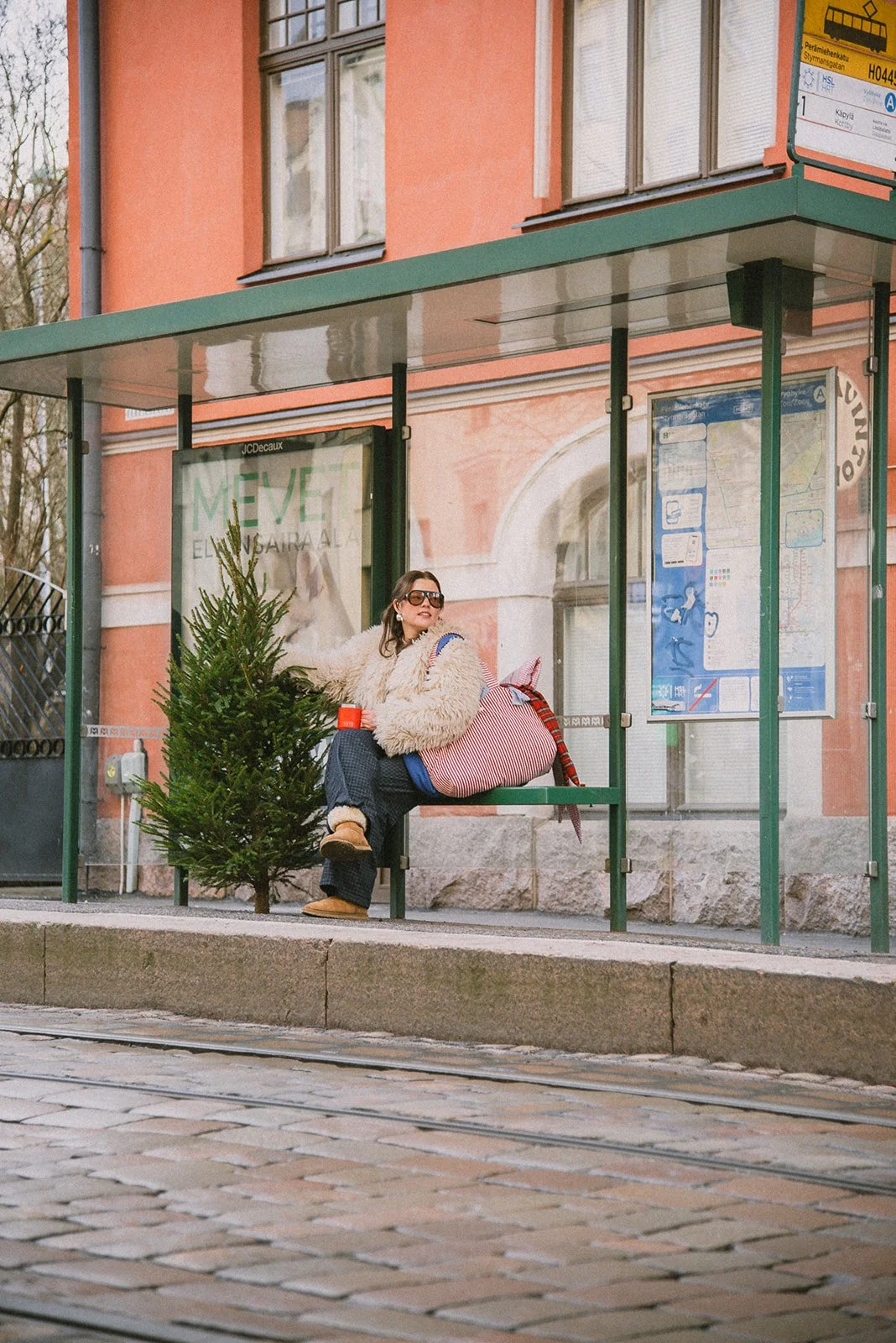 Edlan yrityskuvaus Helsingissä. Yrittäjä istuu ratikkapysäkillä brändin laukut olalla ja pitelee joulukuusta pystyssä.