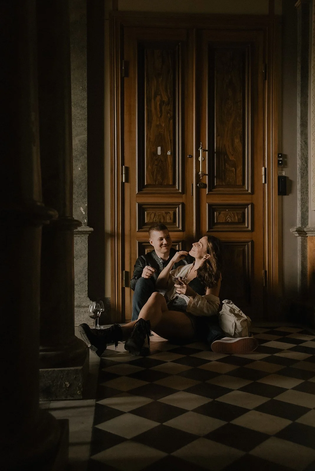 Pariskunta istuu lattialla suuren puisen oven edessä, hymyilevät ja juttelevat. Pariskuntakuvaus Helsinki.