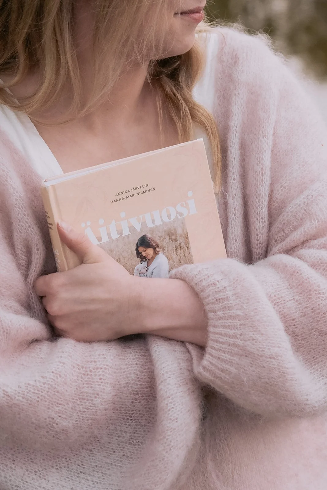 Brändikuva, jossa yrittäjän halaa kirjaa sylissään, päällä vaaleanpunainen neule.