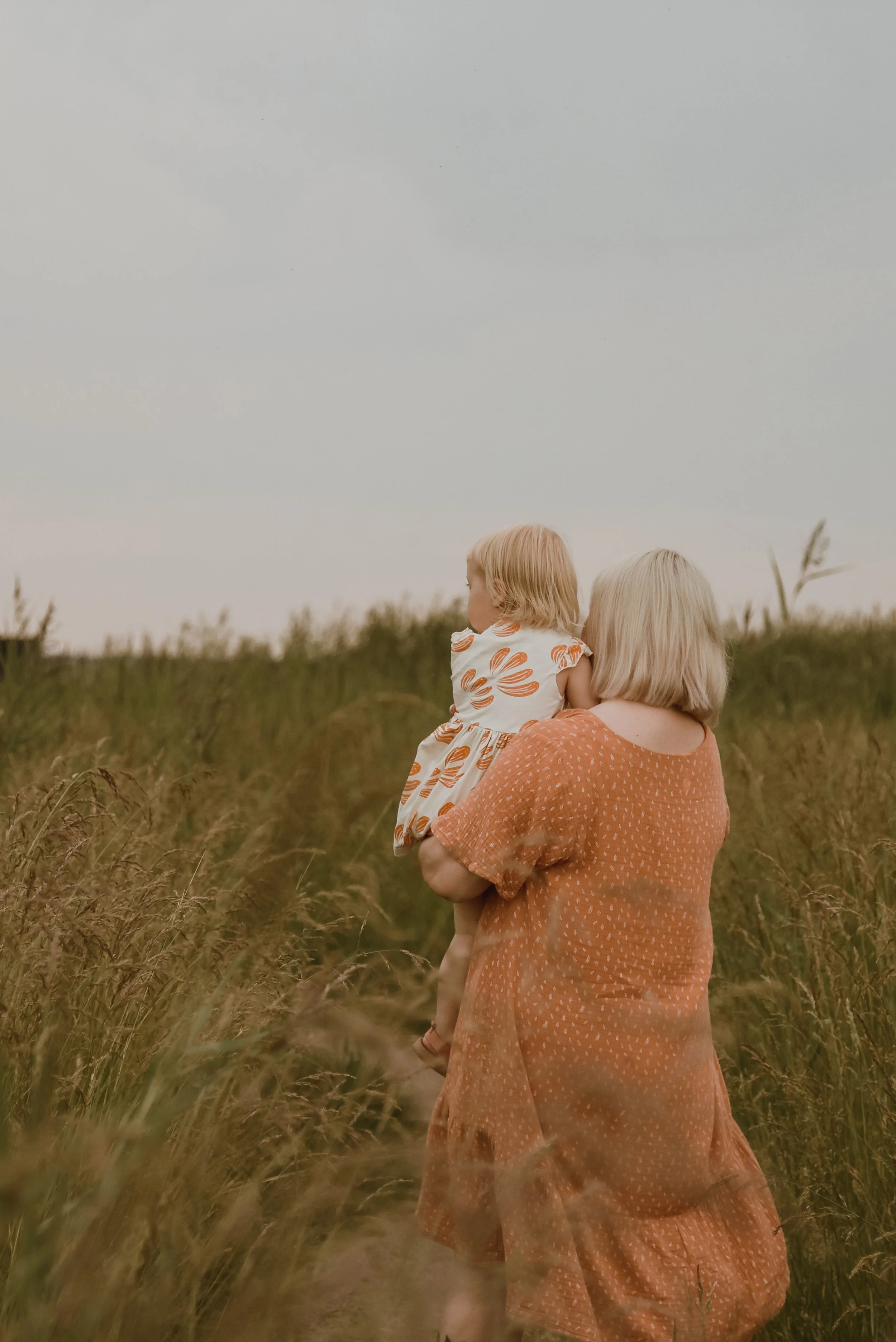 Perhekuvaus luontomaisemassa, äiti kantaa pientä lasta sylissään heinikossa. Kuvannut perhekuvaaja Heidi Väisänen.