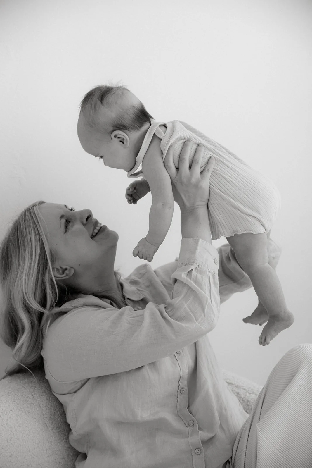 Äiti nostaa vauvaa ilmaan ja hymyilee iloisesti. Äiti-lapsikuvaus Heidi Väisäsen studiolla.