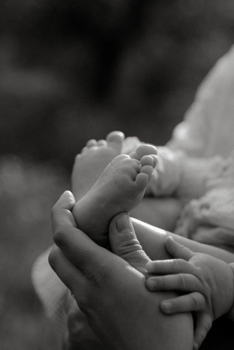 Mustavalkokuva vauvan varpaista ja sormista, joita aikuisen käsi pitelee.