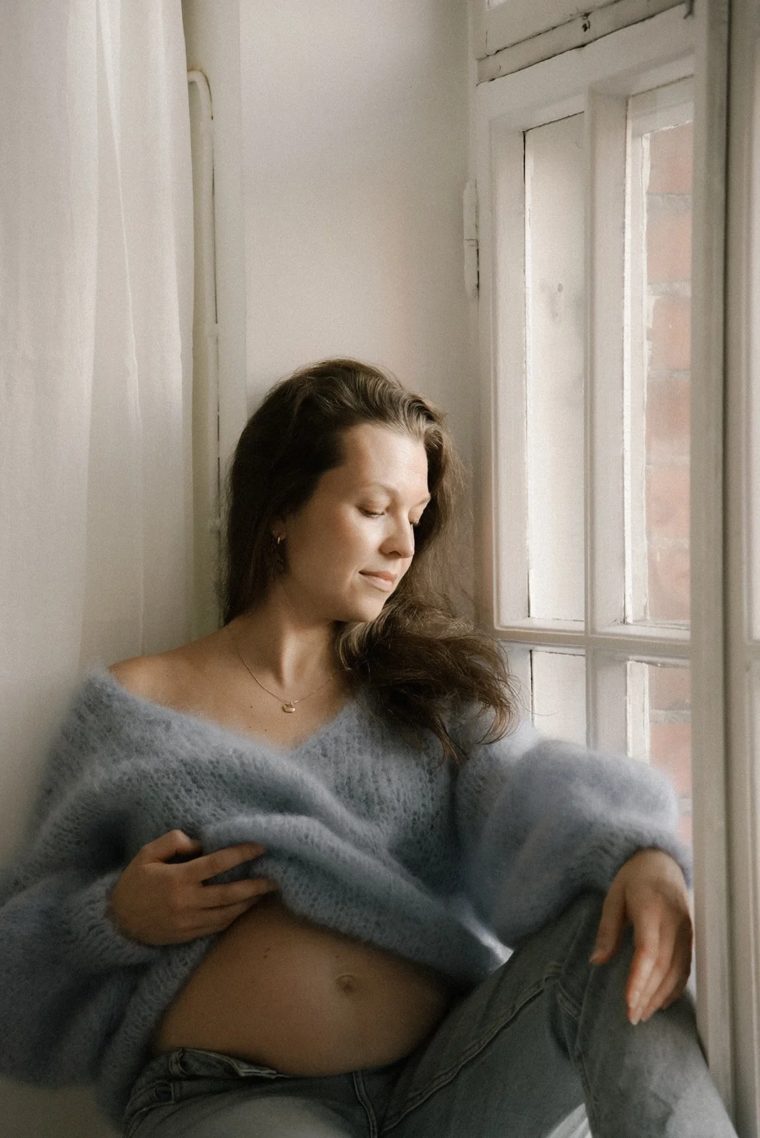 Odottava äiti istuu ikkunan edessä vaaleansininen neule yllään. Kuvan on ottanut raskausajankuvaaja Heidi Väisänen.