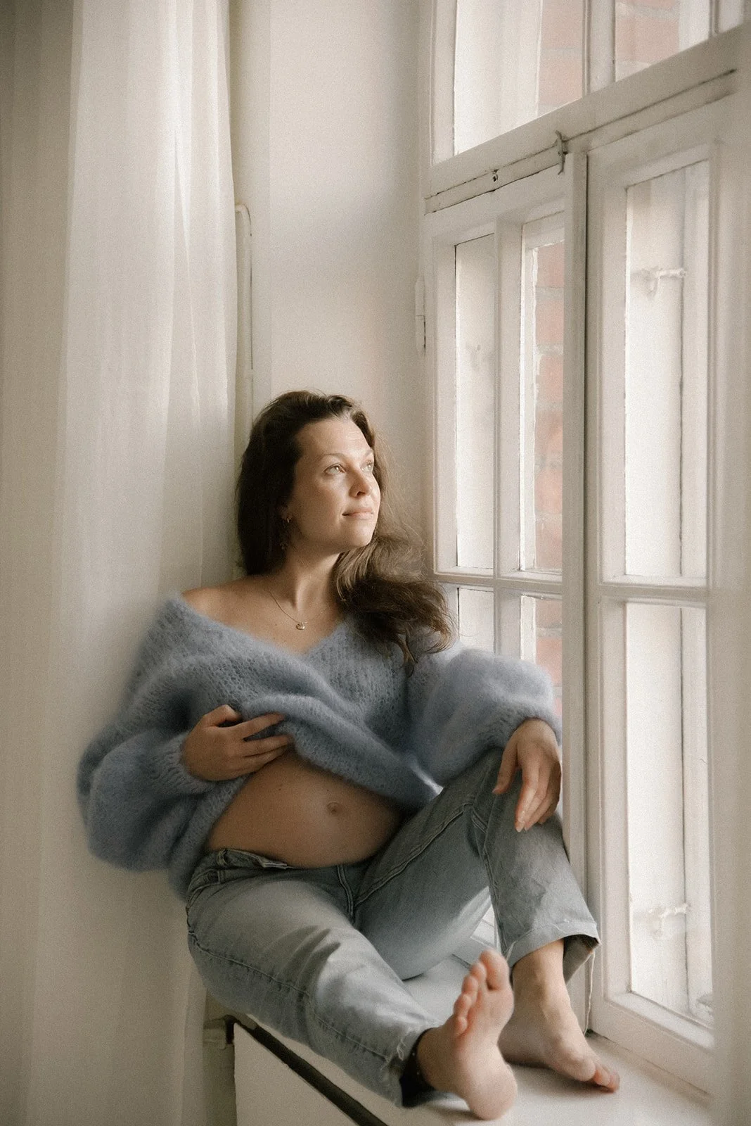 Odottava äiti istumassa ison ikkunan edessä. Odotuskuvaus Heidi Väisäsen studiolla Keravan Saviossa.