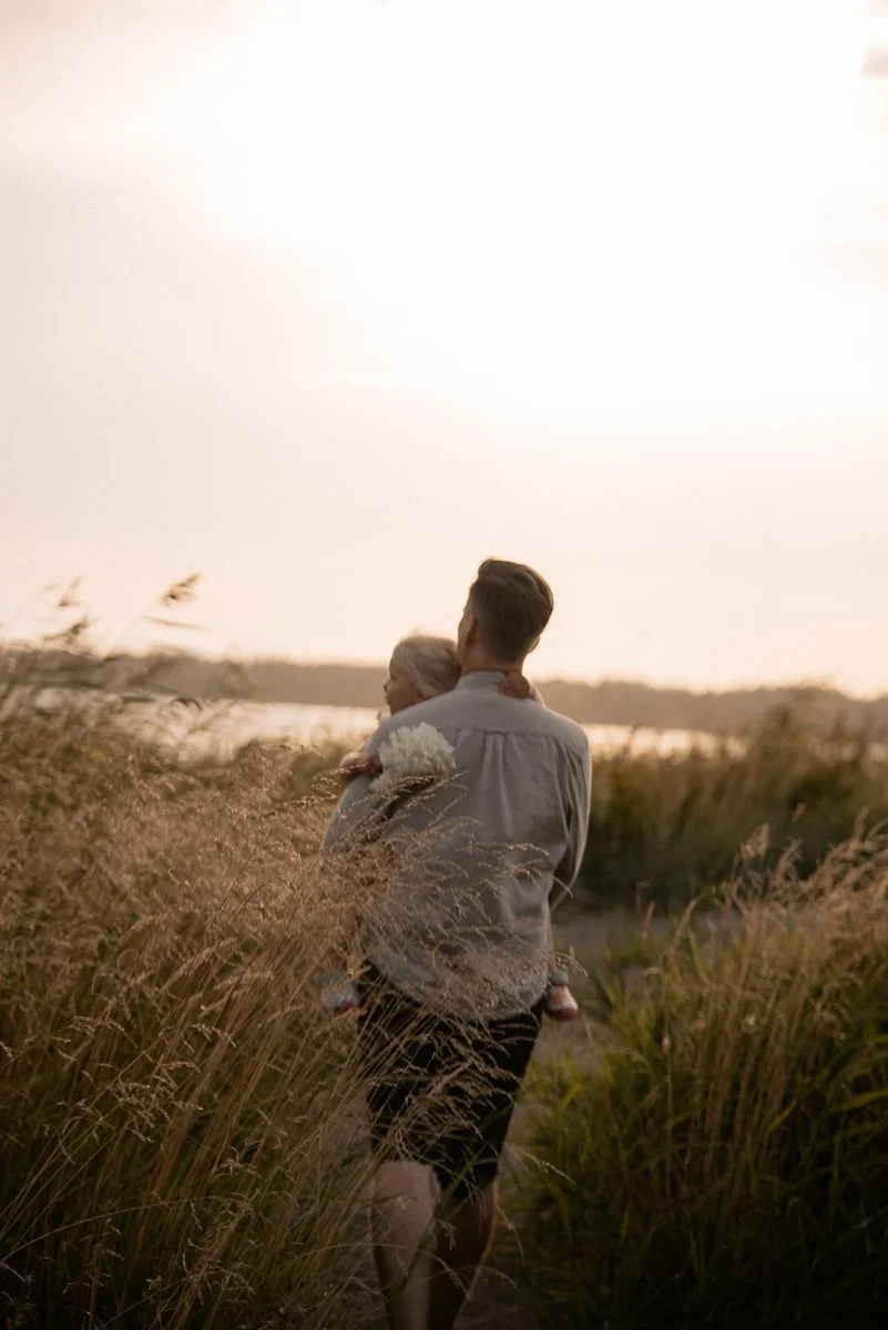 Perhekuvaus miljöössä, isä kantaa lasta korkeassa heinikossa ilta-auringossa. Kuvannut valokuvaaja Heidi Väisänen.