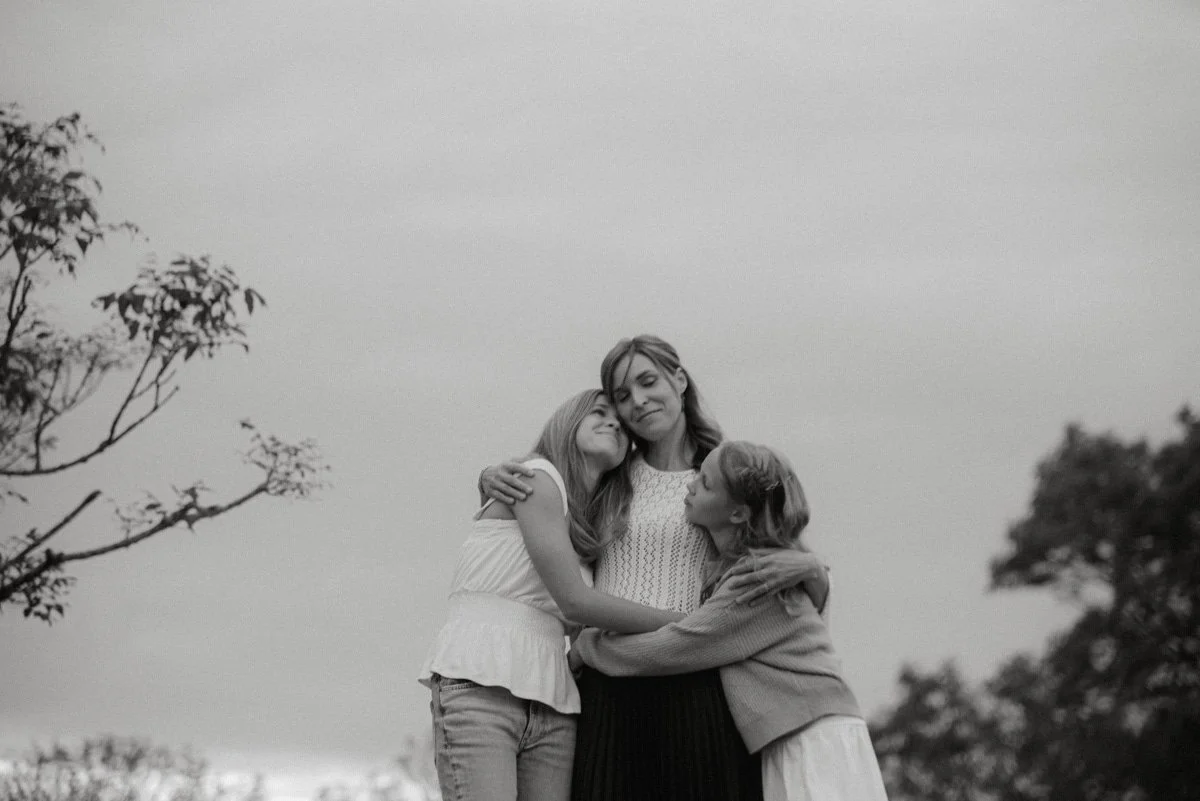 Perhekuvaus Helsinki, äiti ja tyttäret halaavat toisiaan luontomaisemassa.