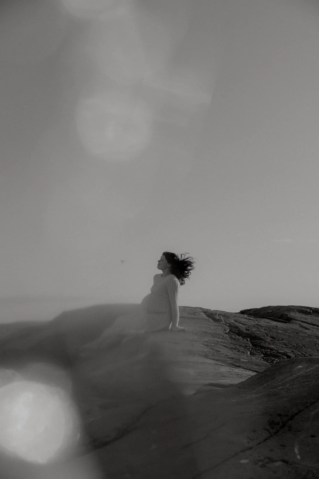 Mustavalkoinen kuva, jossa odottavan äidin hiukset hulmuavat tuulessa. Raskauskuvaus luontoympäristössä.