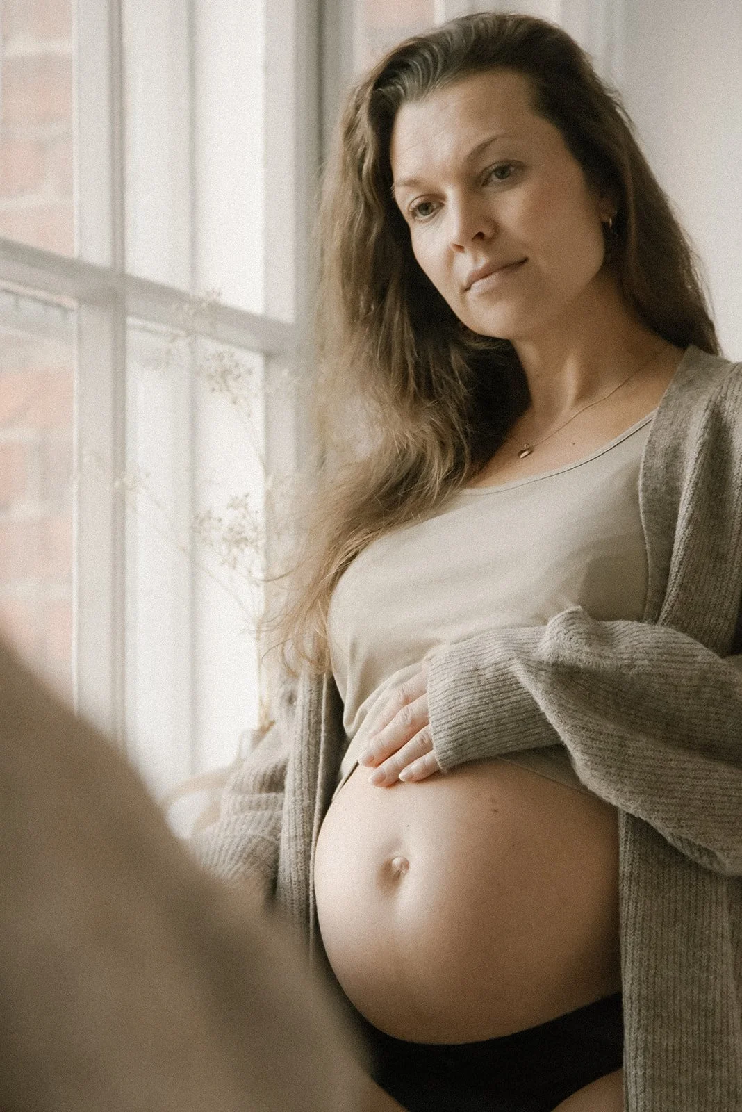 Raskaana oleva nainen ikkunan edessä lempeässä luonnonvalossa. Raskauskuvaus studiolla.