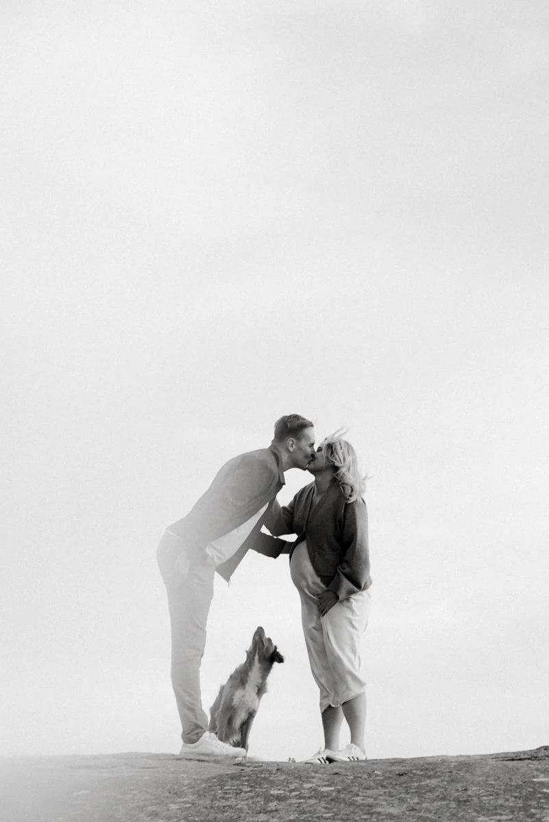 Mustavalkoinen kuva, jossa odottava äiti ja mies suutelevat toisiaan, maassa istuu koira.