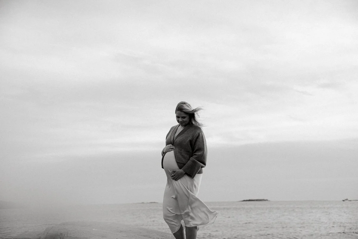 Mustavalkoinen kuva odottavasta äidistä mekossa ja neuleessa.