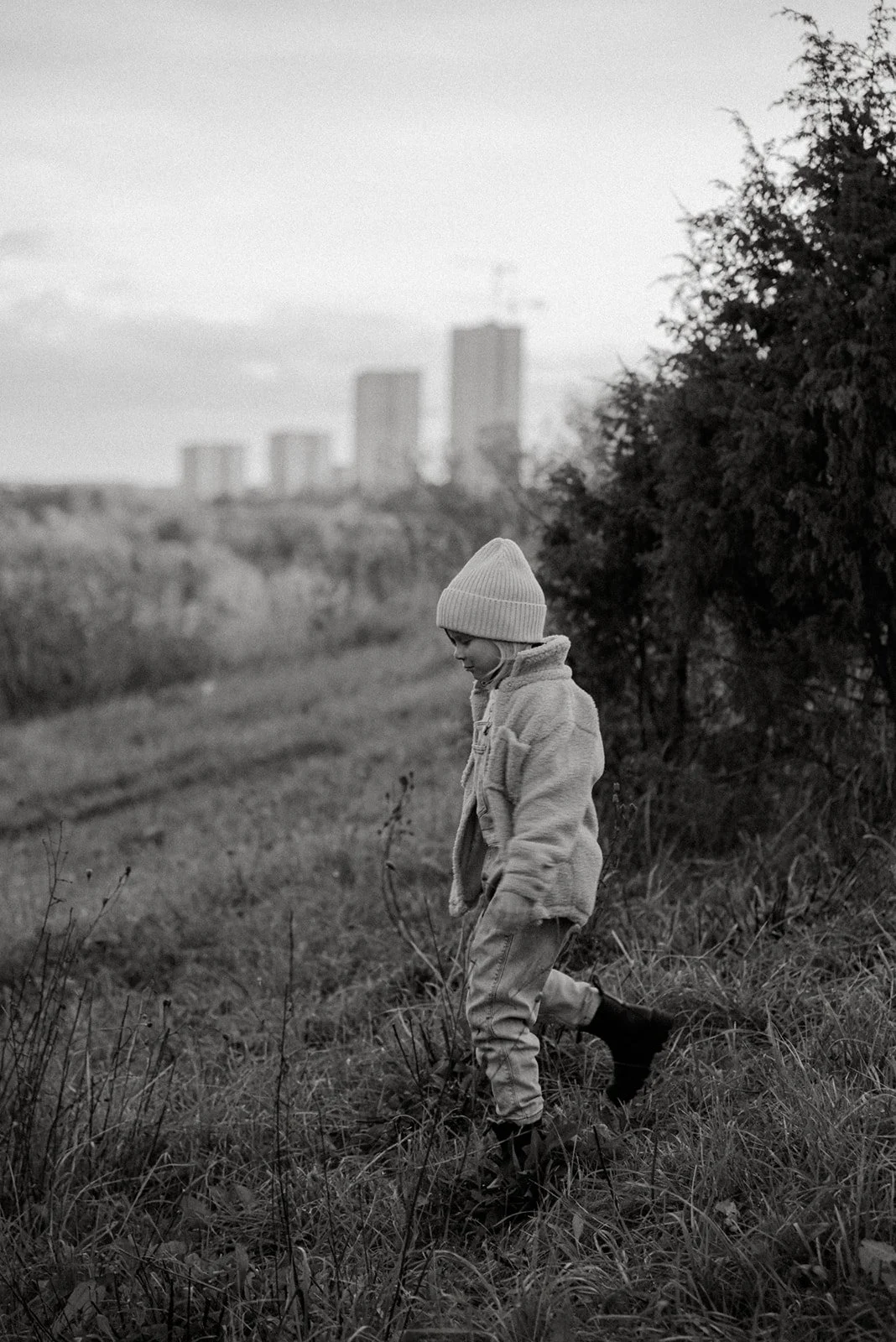 Mustavalkokuva pienestä lapsesta, joka kävelee luonnossa, taustalla kaupunkimaisema.