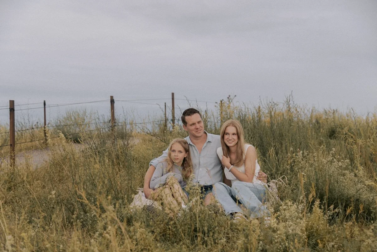 Perhekuvaus luontomaisemassa, isä ja kaksi lasta istuvat heinikössä. Kuvannut perhekuvaaja Heidi Väisänen.