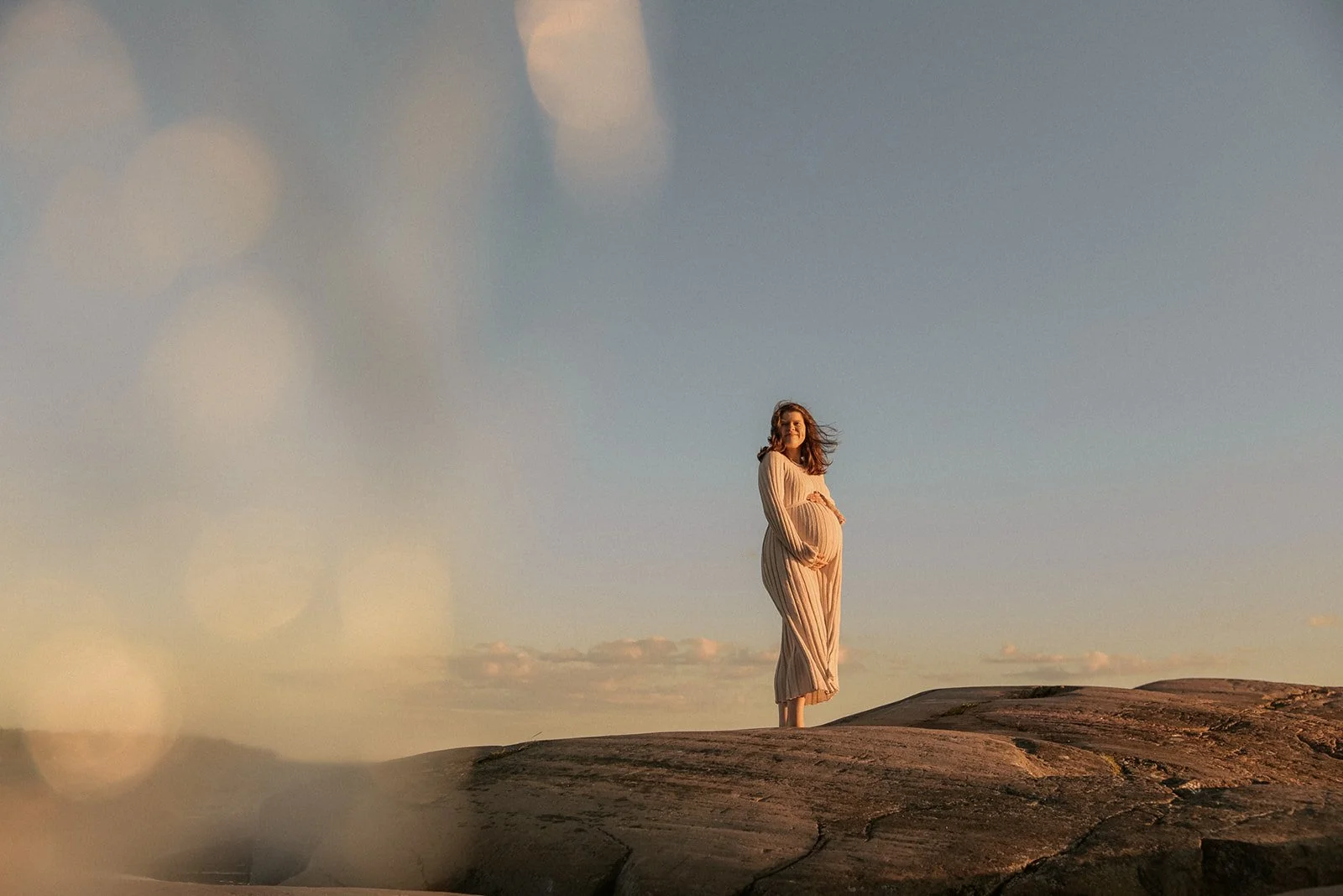 Odottava äiti vaaleassa mekossa kaunista sinistä taivasta vasten.