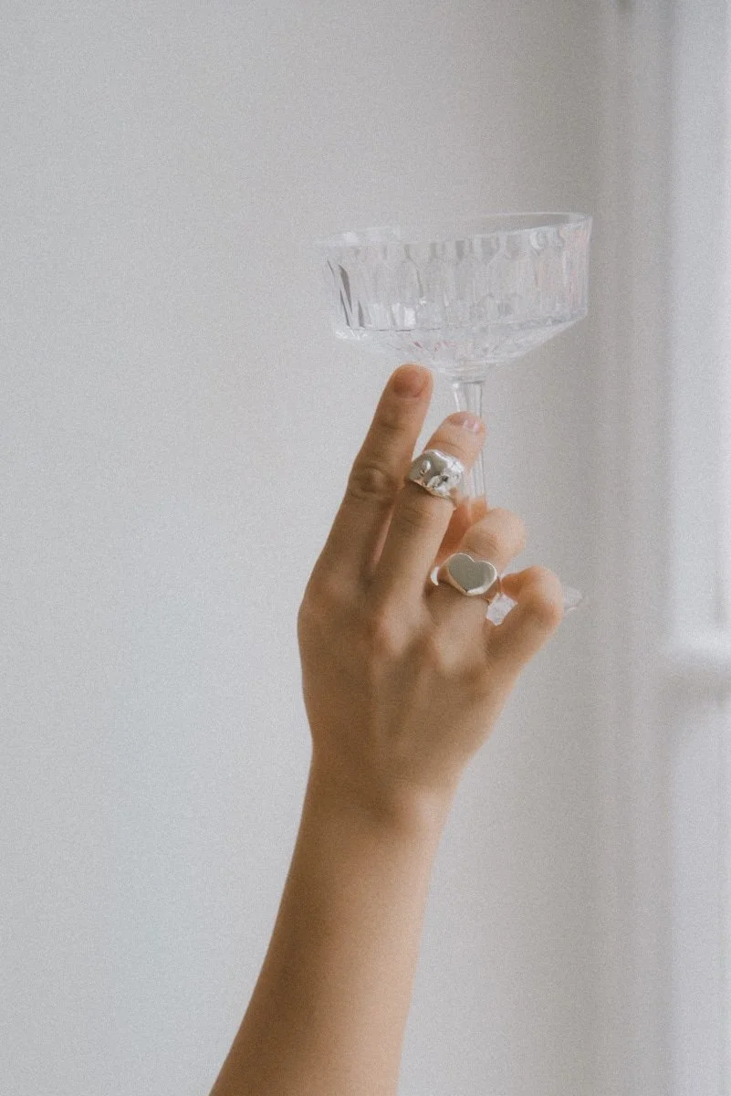 Minimalistinen tuotekuva sormuksista, jossa käsi pitelee lasia.