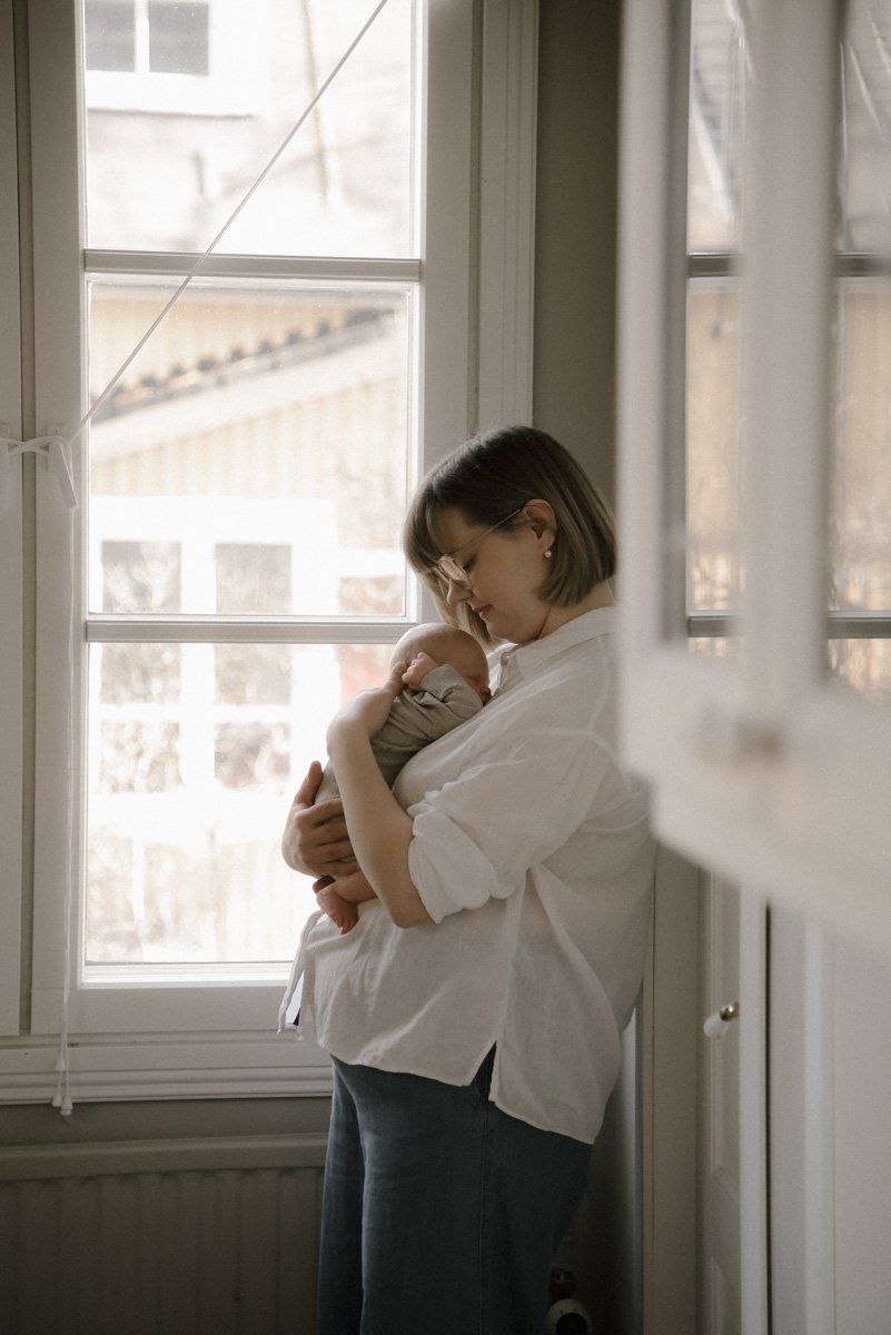 Äiti seisoo valoisan ikkunan edessä lapsi sylissään. Perhekuva Helsinki.