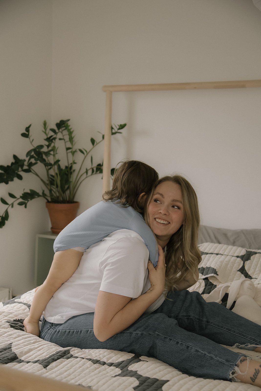 Äidin reppuselässä lapsi sängyllä. Perhekuvaus kotona.