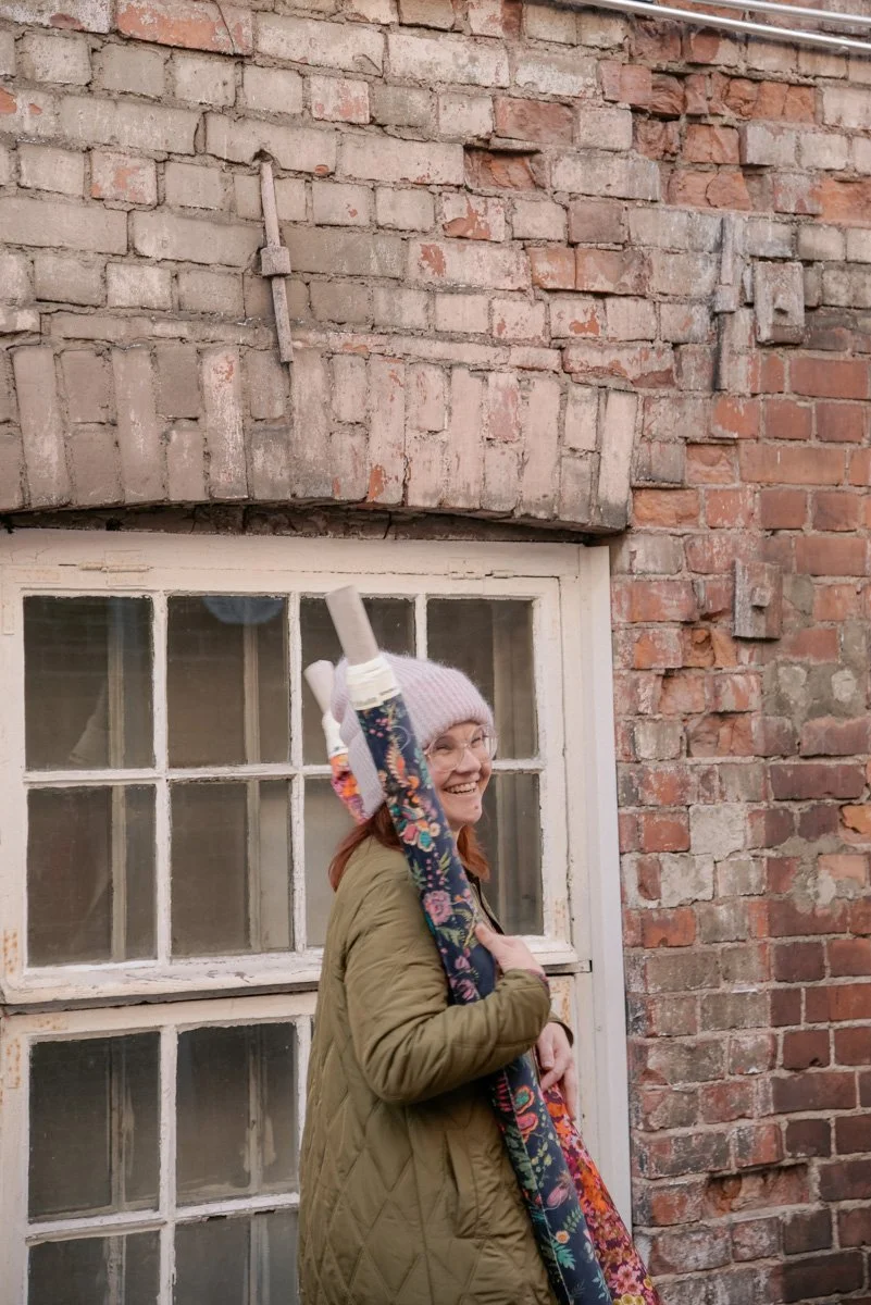 Verhoomo Vanhan Viehätys yrityskuvaus, yrittäjä Virve Taljavirta kävelee ulkona tiilisen talon vieressä ja kantaa käsissään kangasrullia.