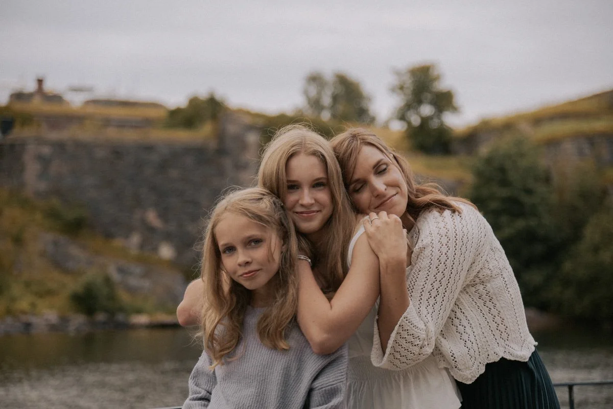 Perhekuvaus miljöössä, äiti ja tyttäret halaavat toisiaan. Kuvannut perhekuvaaja Heidi Väisänen.