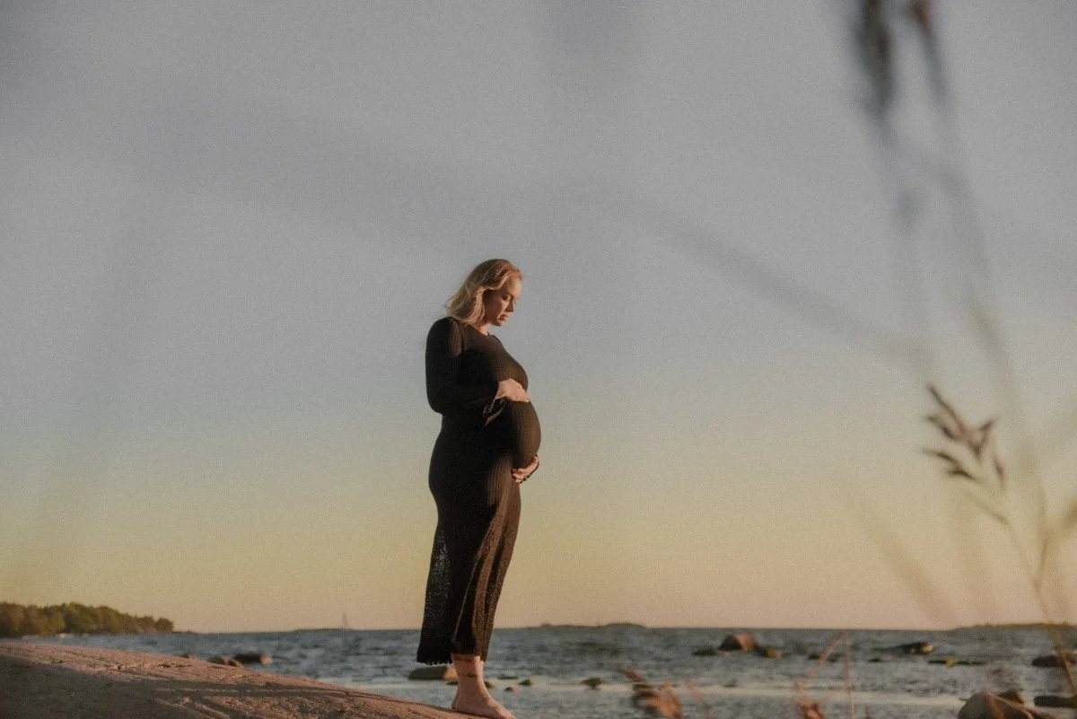 Lasta odottava nainen pitelee vatsaansa ja seisoo meren äärellä ilta-auringossa. Kuvannut Heidi Väisänen, joka valokuvaa odottavia äitejä.