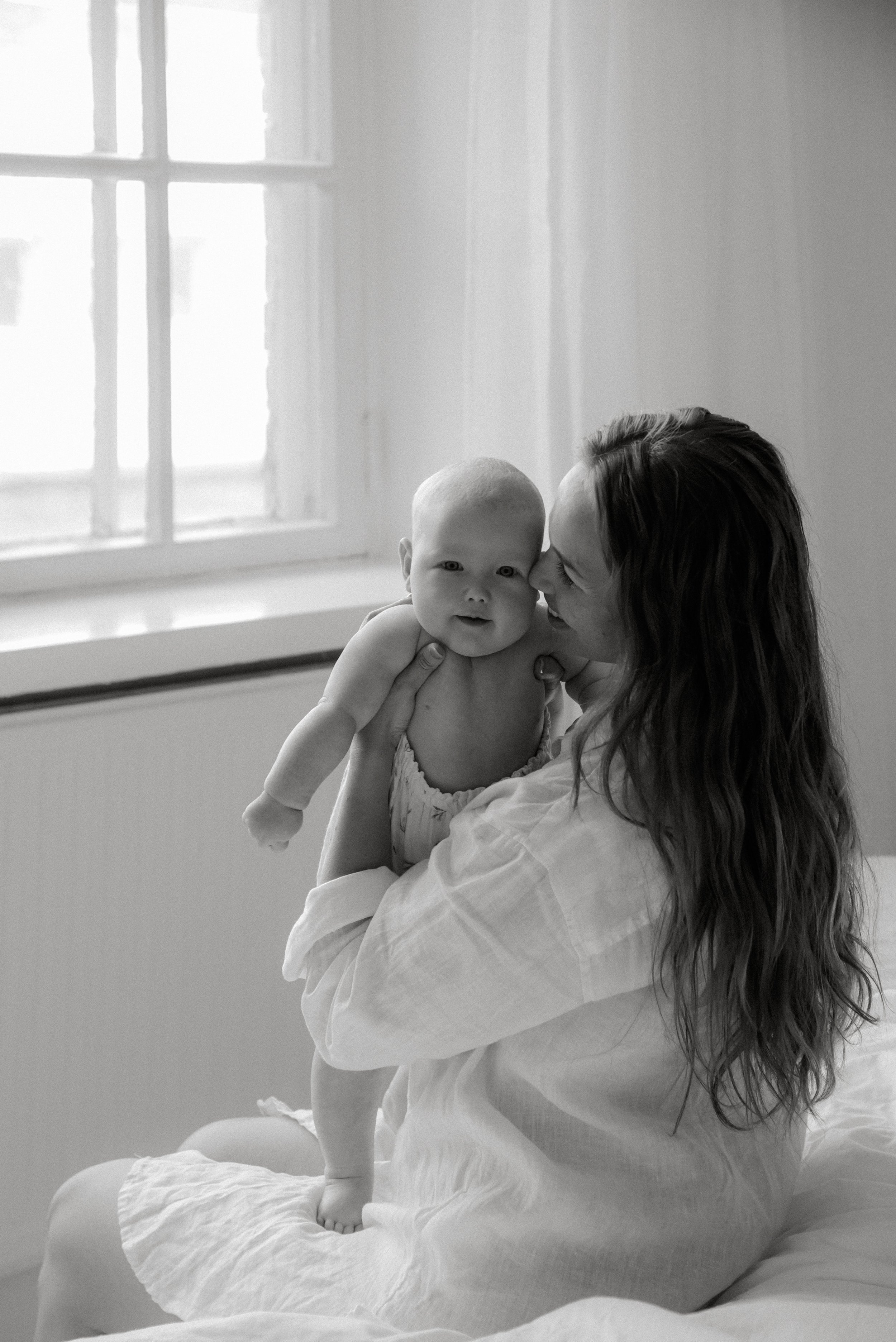 Äiti pitää vauvaa sylissään Heidi Väisäsen valoisan studion ikkunan edessä, molemmat hymyilevät. 