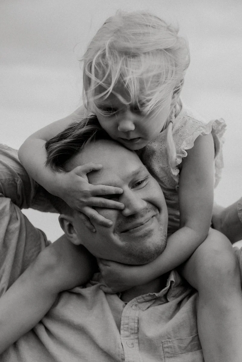 Mustavalkoinen perhekuva, jossa pieni lapsi istuu isän olkapäillä.