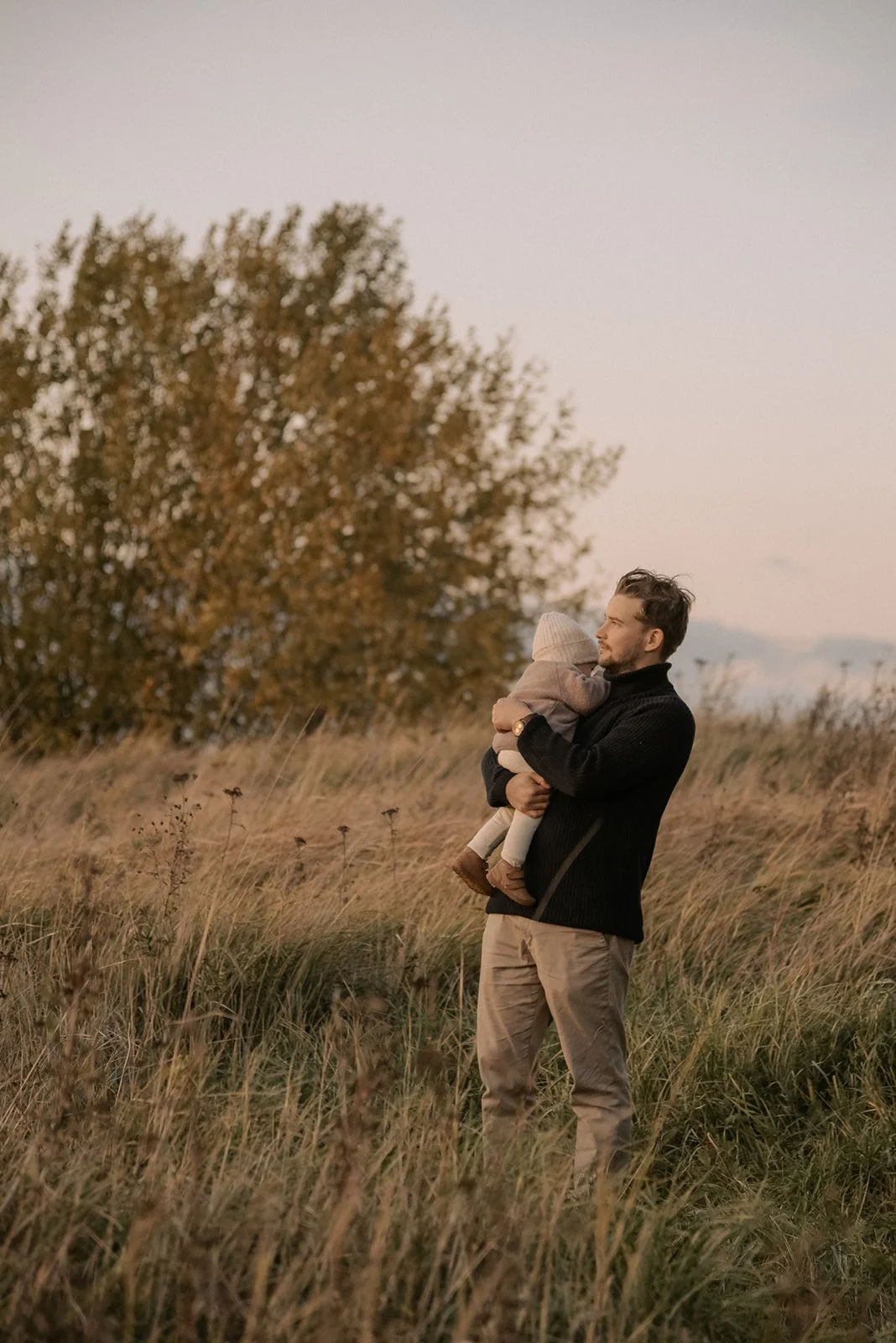 Isä seisoo heinikossa lapsi sylissään. Perhekuvaus Espoo.