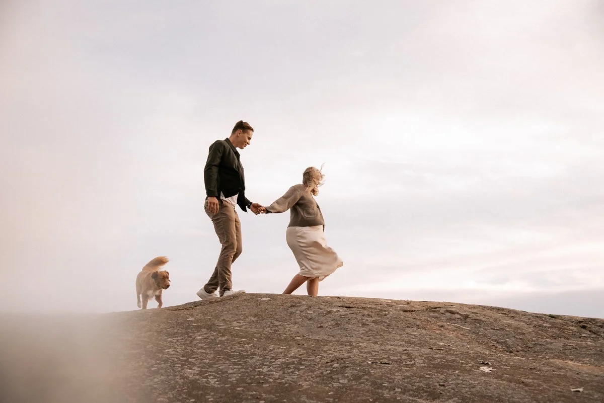Käsi kädessä kävelevä pariskunta ja koira kallioilla. Raskauskuvaus luonnossa.