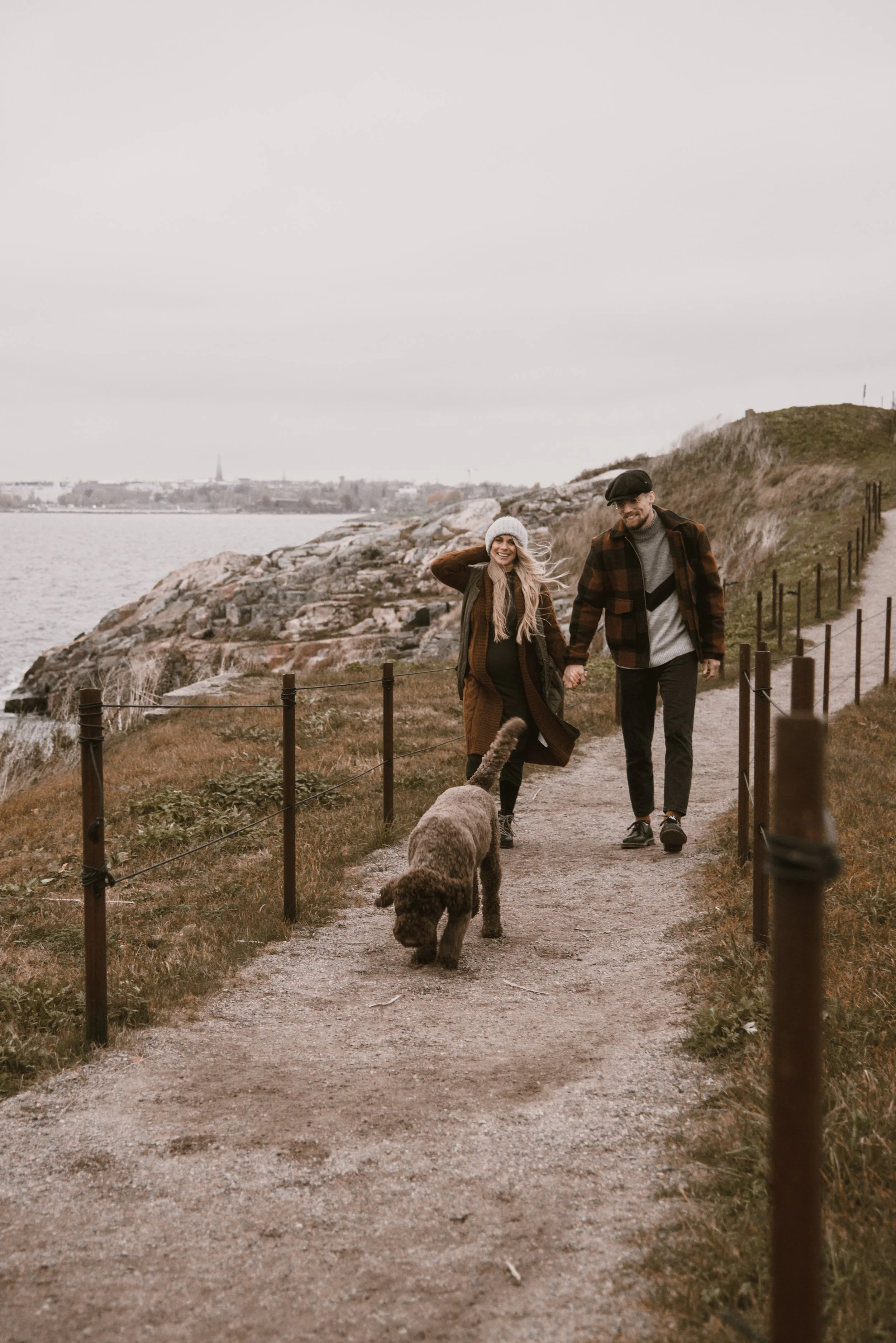Pariskunta ja koira kävelee polkua pitkin. Odotusajan kuvaus miljöössä.