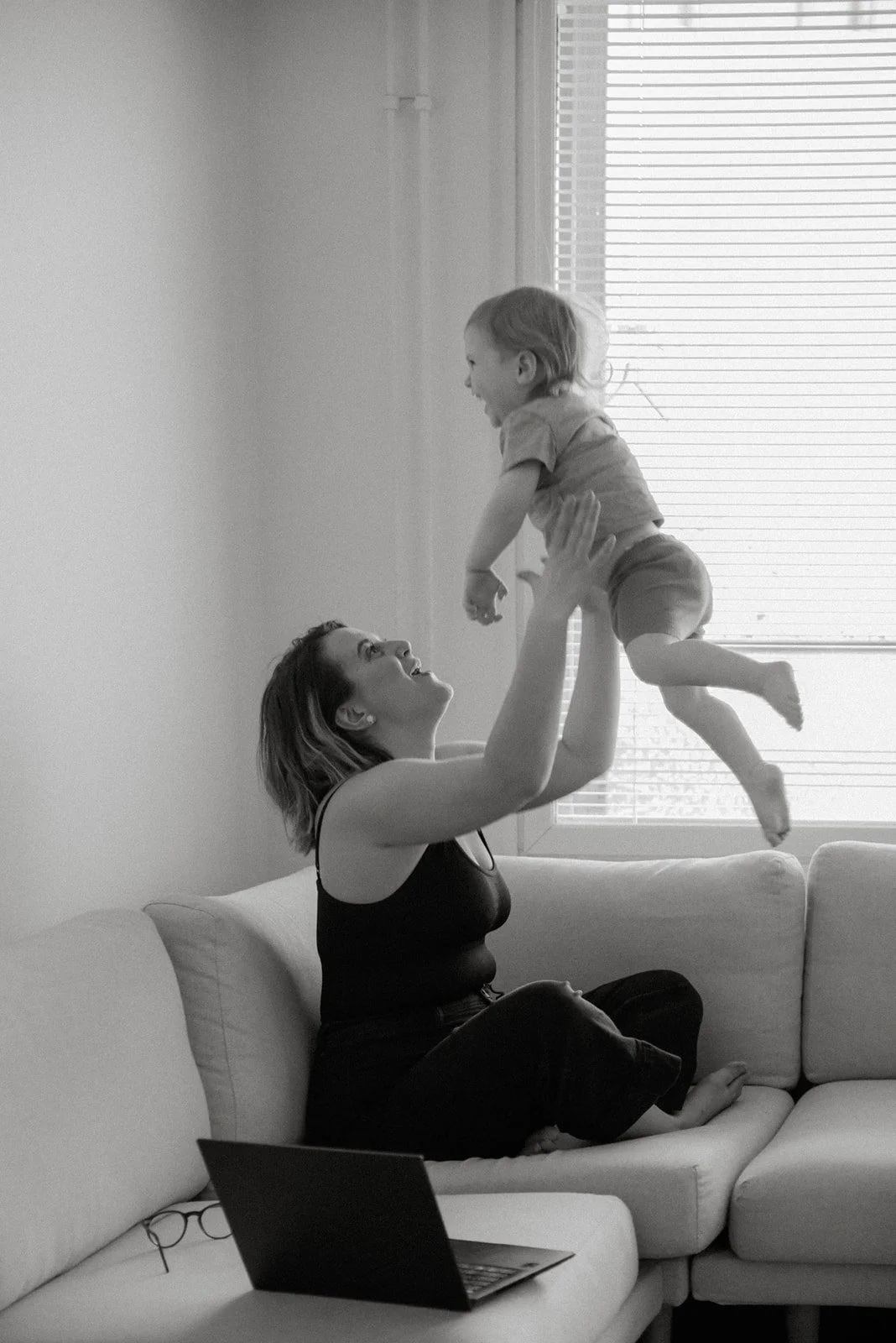 Nainen nostaa lastaan iloisesti käsivarsilleen. Perhekuvaus kotona.