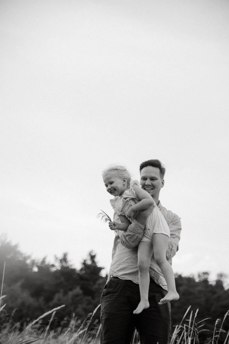 Perhekuvaus Espoo, pieni nauravainen lapsi isän sylissä luontomaisemassa.