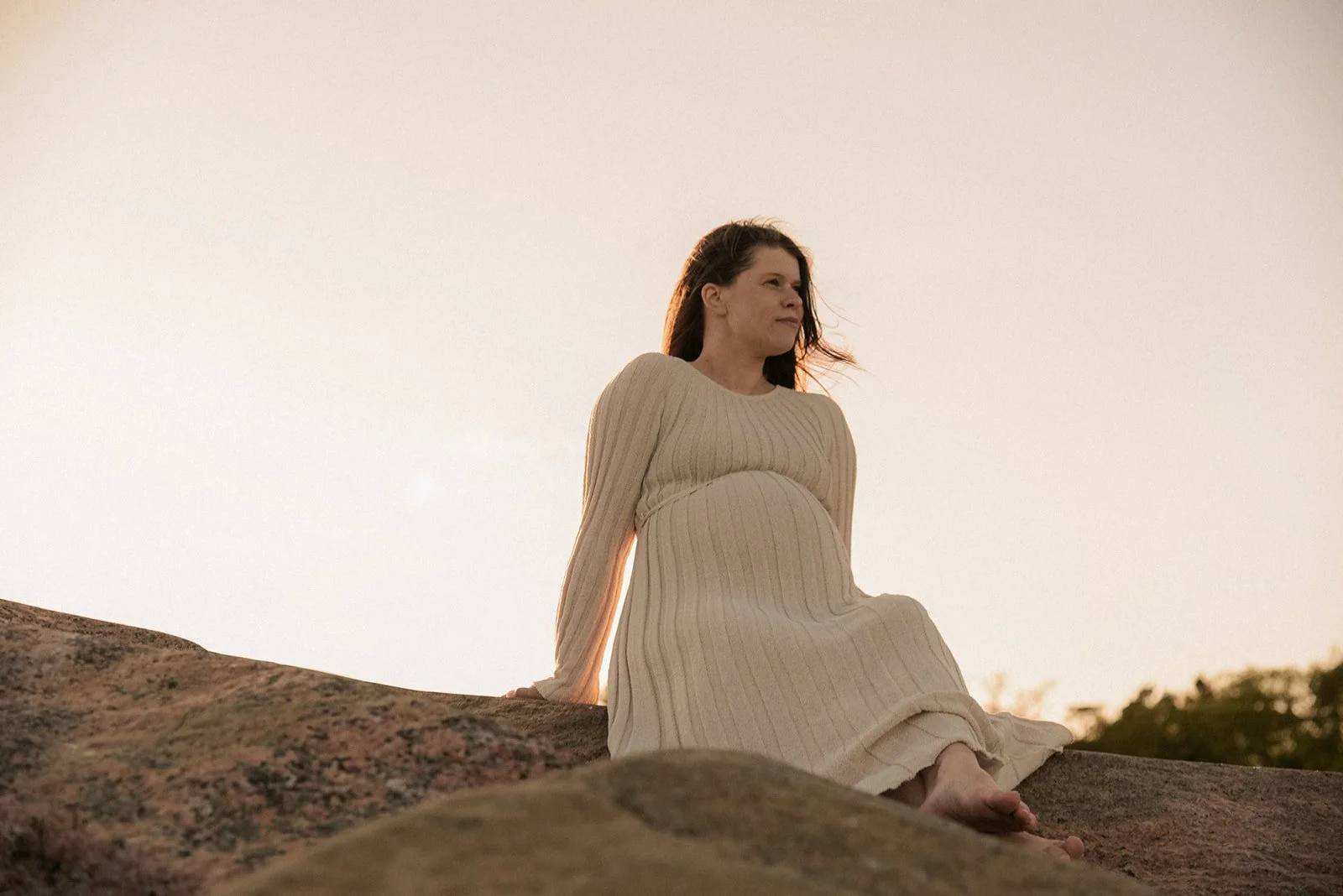 Kalliolla istuva odottava äiti vaaleassa mekossa. Odotusajankuvaus miljöössä.