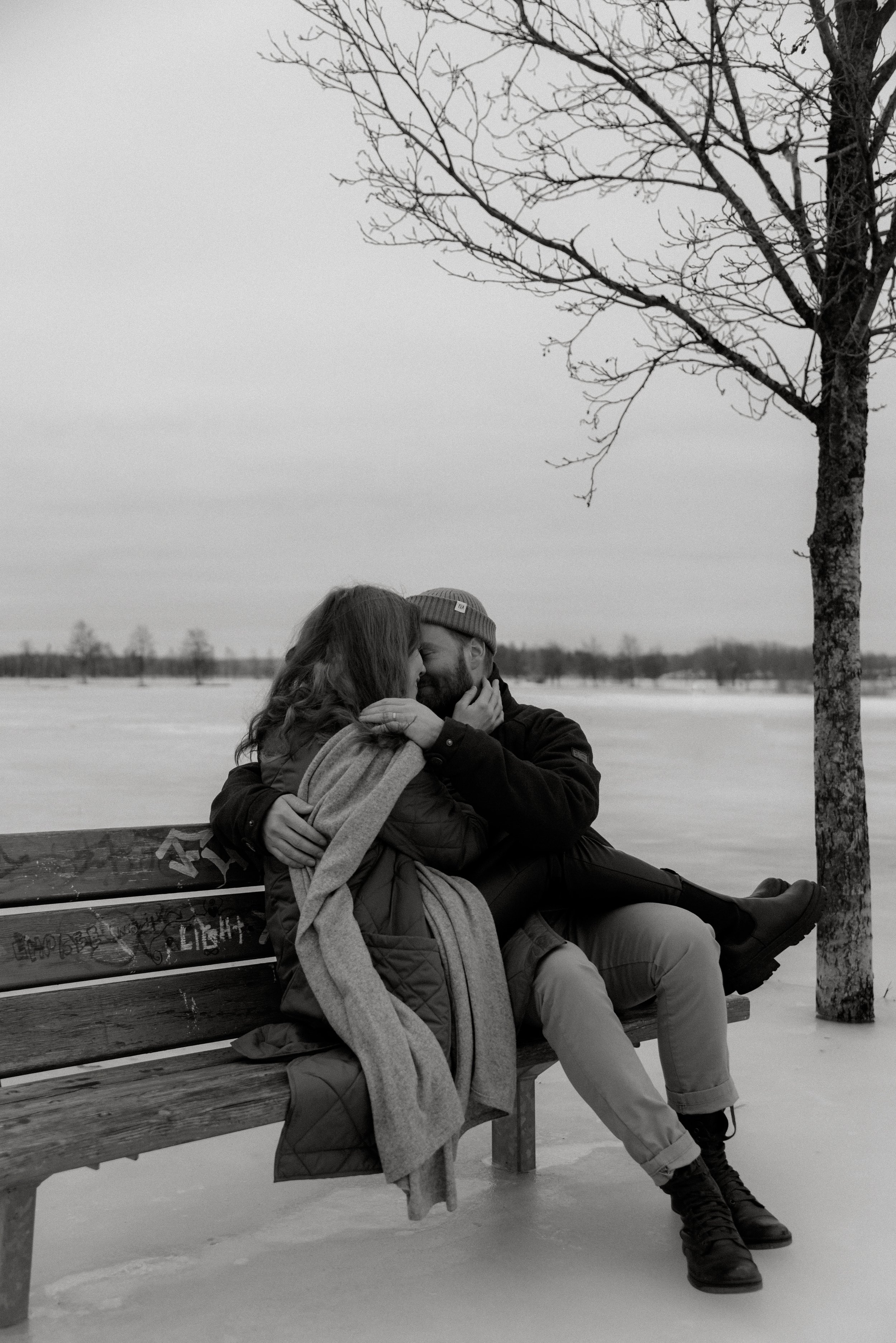Mustavalkoinen pariskuntakuva, nainen ja mies halaavat puiston penkillä.