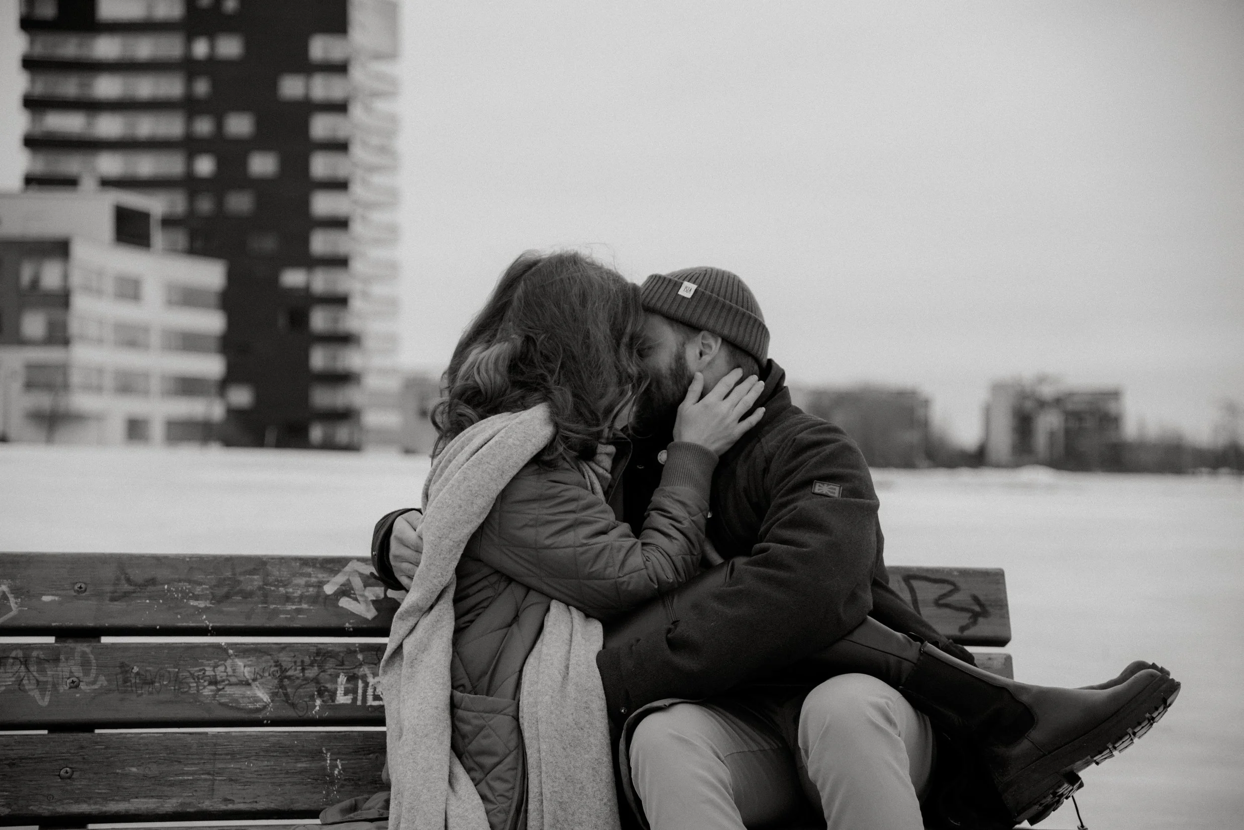 Pariskunta suutelee toisiaan ulkona penkillä, taustalla rakennuksia. Pariskuntakuvaus Espoo.