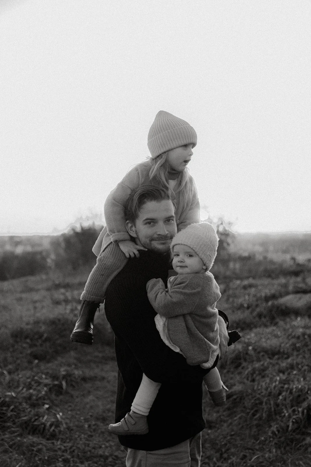 Perhekuvaus miljöössä, isä kantaa kahta pientä lasta, toista olkapäillä ja toista sylissä. Kuvannut perhekuvaaja Heidi Väisänen.