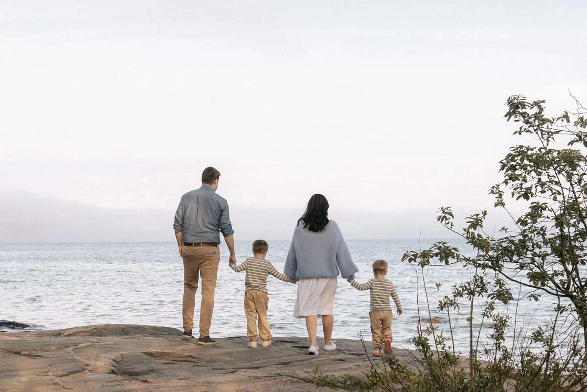 Nelihenkinen perhe rantakallioilla meren äärellä, pitävät toisiaan käsistä kiinni.