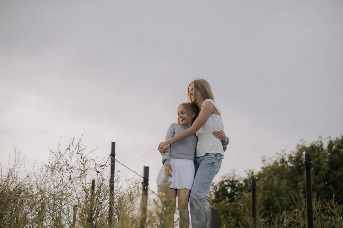 Perhekuvaus Espoo. Äiti ja lapsi halaavat iloisesti ulkona, taustalla pensaikko ja harmaa taivas.