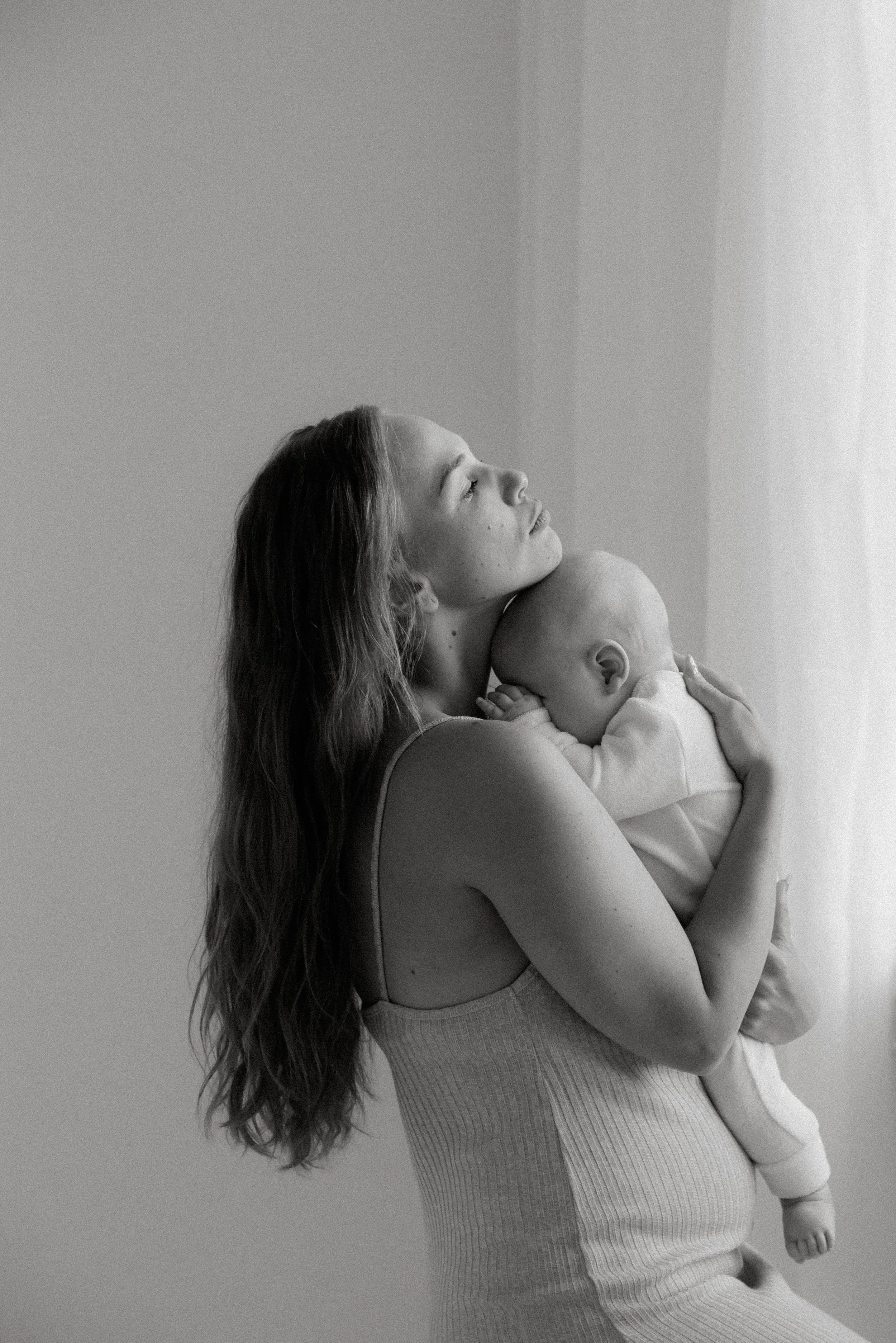 Äiti-lapsikuvaus, äiti pitelee lasta sylissään kasvot kohti valoa tulvivaa ikkunaa.