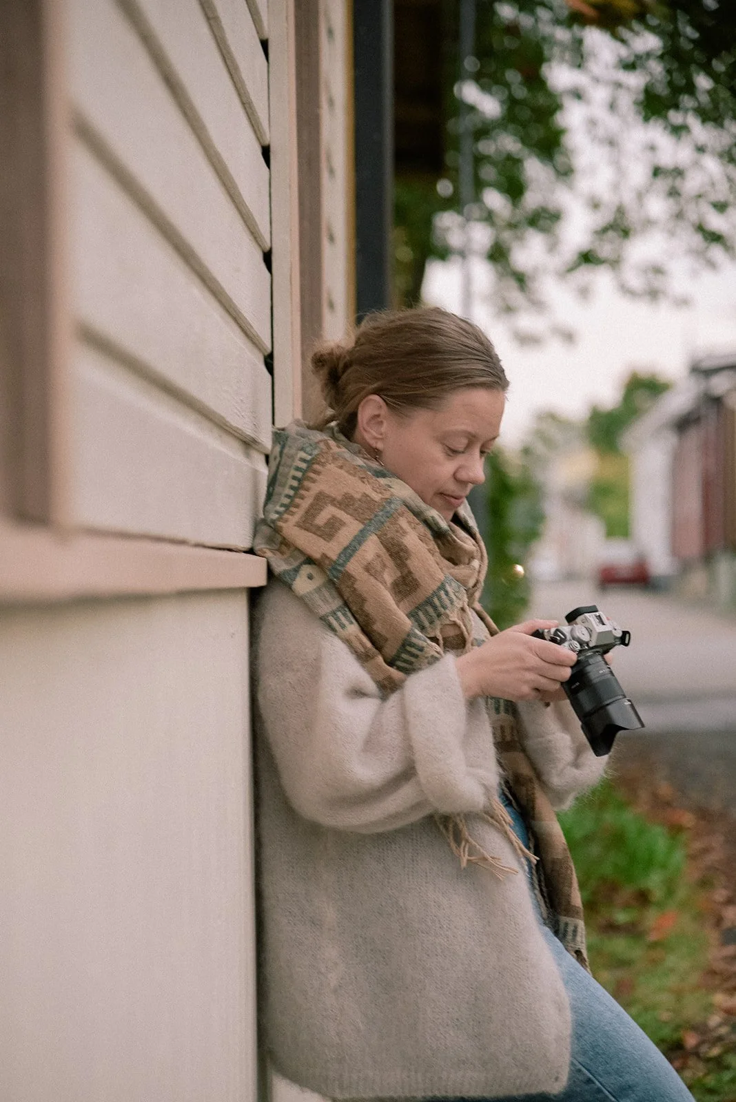 Yrityskuvaus idyllisessä puutalomaisemassa. Naisyrittäjä Laura Laakso nojaa talon seinään ja pitelee käsissään kameraa.