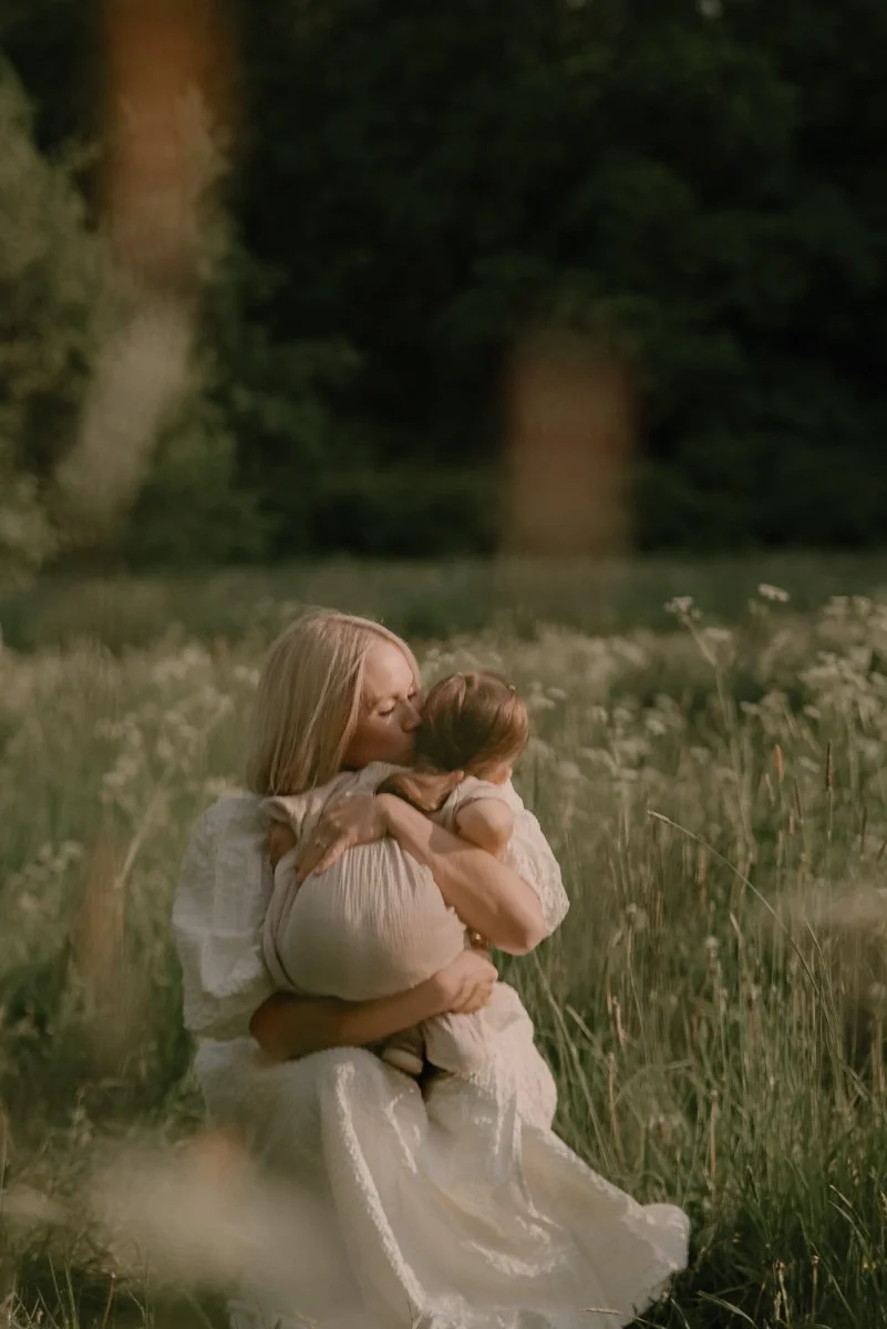 Perhekuvaus ulkona, äiti halaa pientä lasta sylissään heinikossa. Kuvannut valokuvaaja Heidi Väisänen.