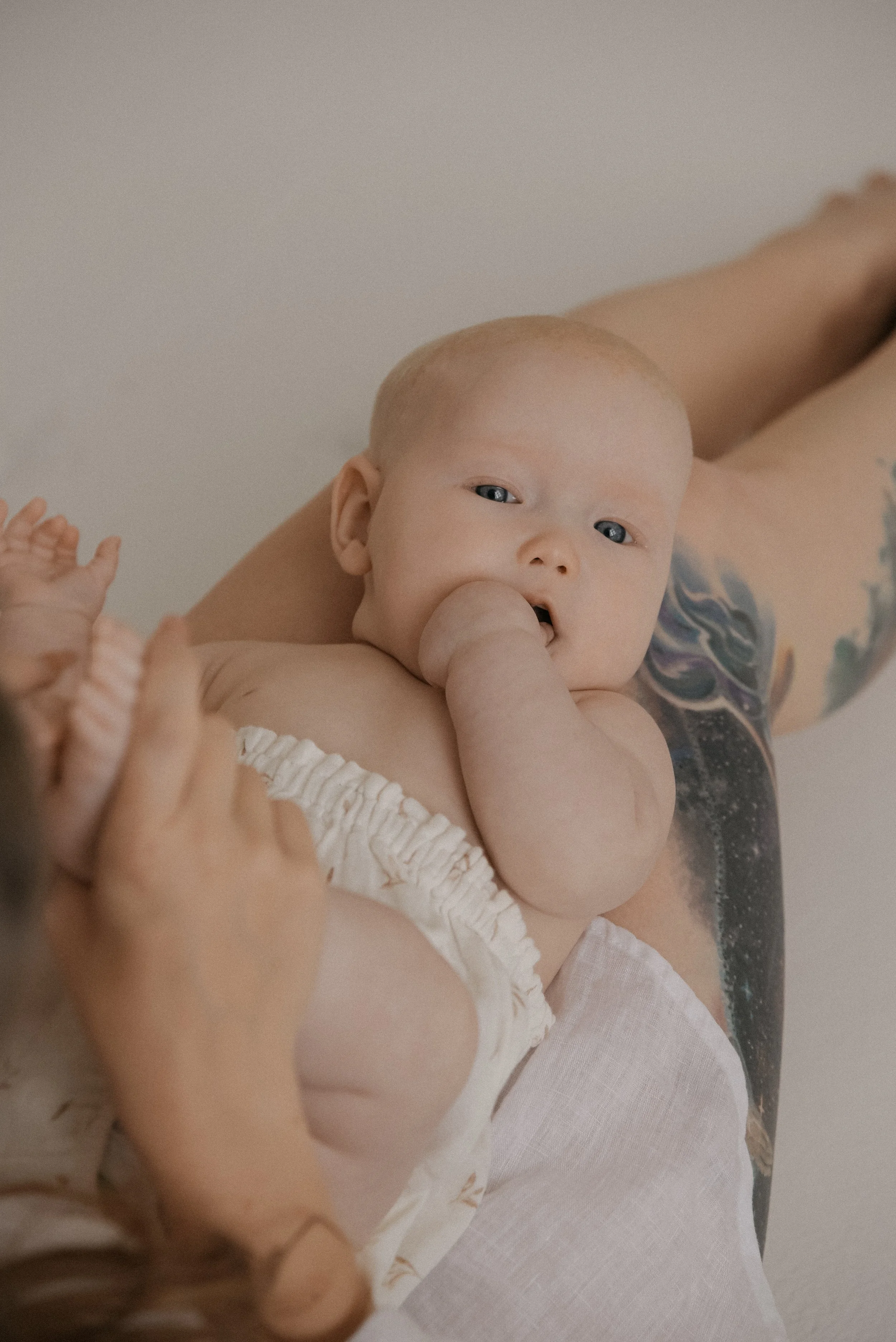 Vauva lepää äidin jalkoja vasten ja mutustaa nyrkkiään. Äiti- ja lapsikuvaus Heidi Väisäsen studiolla.