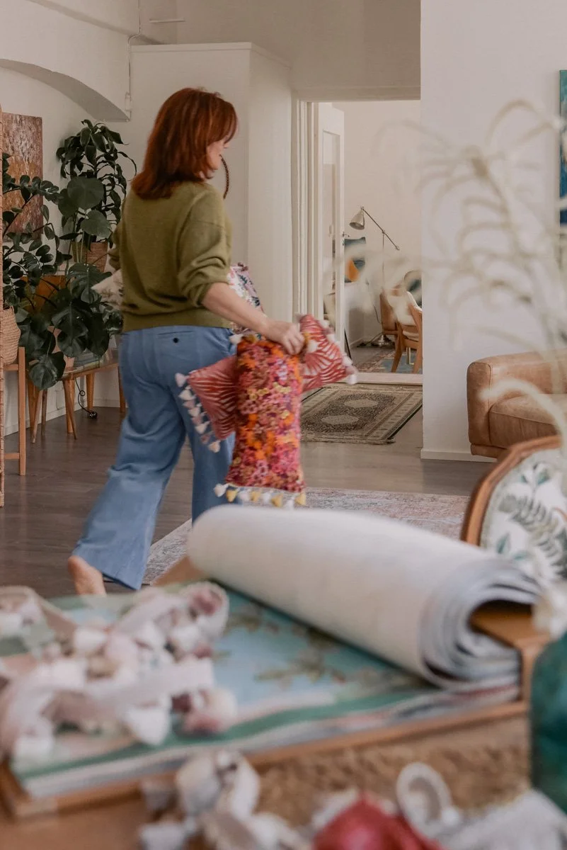 Verhoomoyrittäjä Virve työssään verhoomon tiloissa, kantaa käsissään värikkäitä tyynyjä ja etualalla tarvikkeita. Kuvannut yrityskuvaaja Heidi Väisänen.