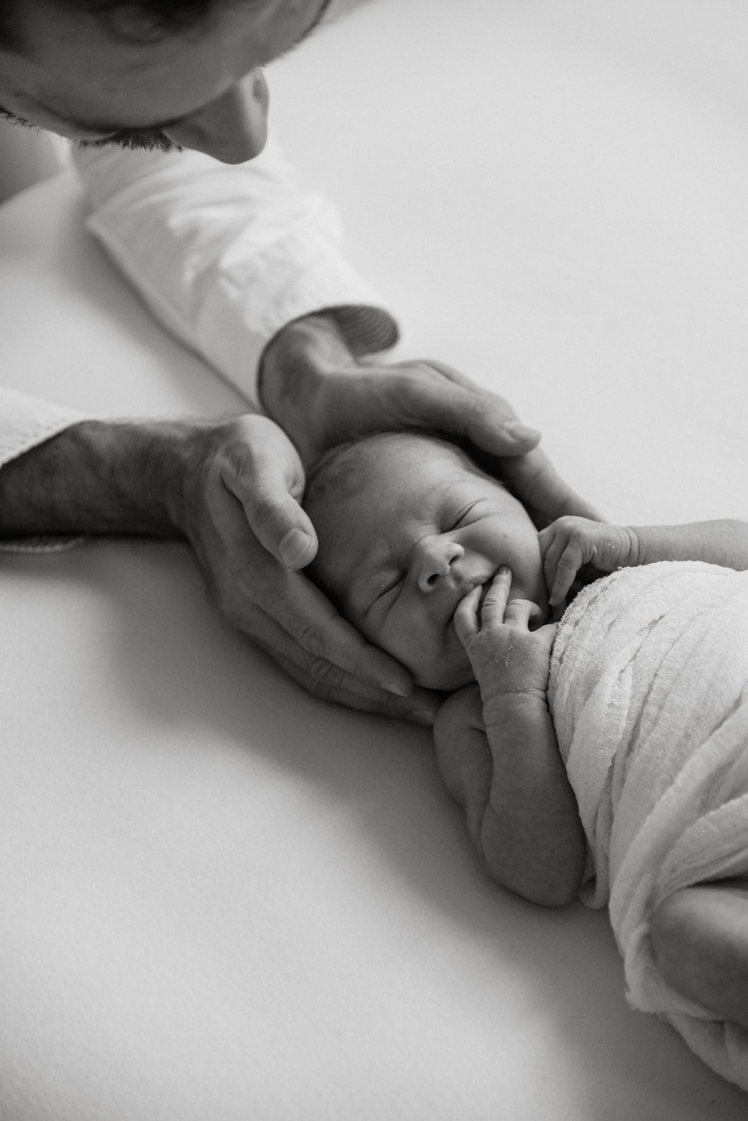 Mustavalkoinen seesteinen kuva, jossa vastasyntynyt vauva nukkuu ja isä pitelee hellästi käsillään vauvan päästä kiinni. Kuvan on ottanut vauvakuvaaja Heidi Väisänen.