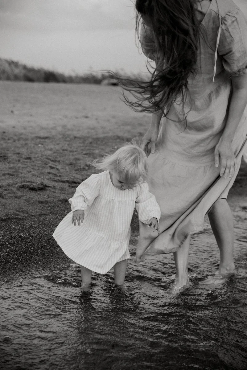 Perhekuvaus meren rannassa, äiti ja pieni lapsi kahlaavat rantavedessä. Kuvannut perhekuvaaja Heidi Väisänen.