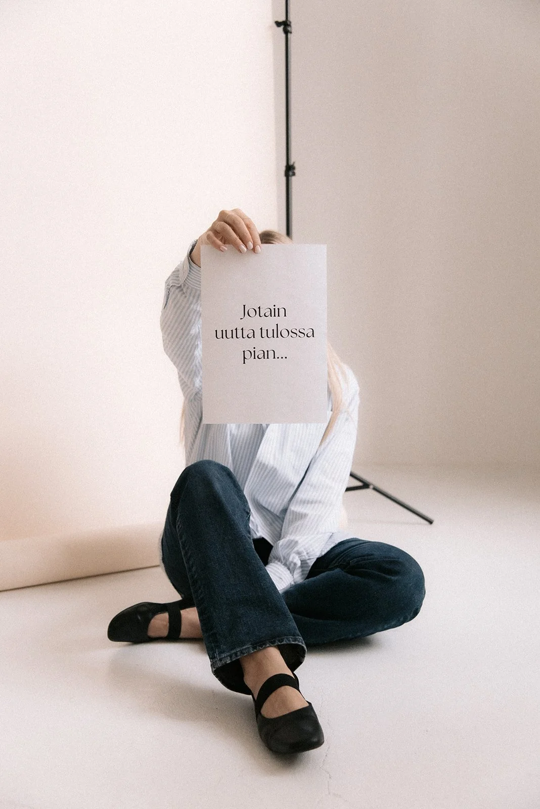 Heartiven yrittäjä istuu valokuvausstudion lattialla ja pitelee paperia, jossa lukee "Jotain uutta tulossa pian...". Kuvannut brändikuvaaja Heidi Väisänen.