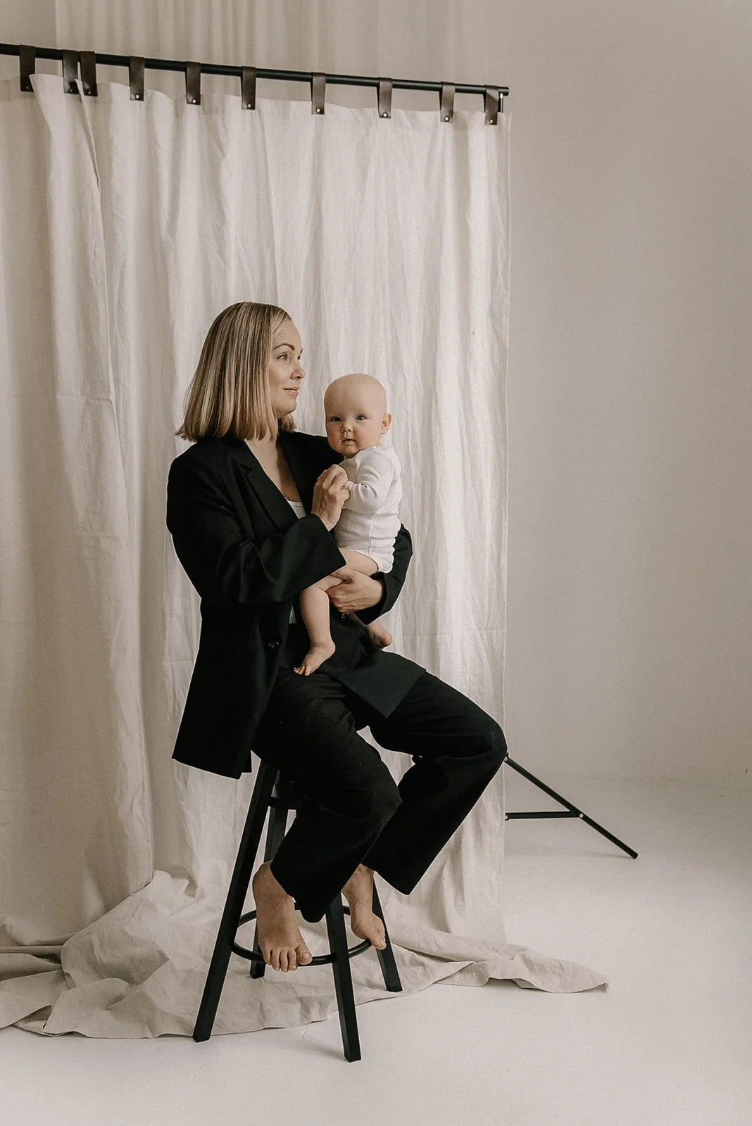 Mustaan jakkupukuun pukeutunut äiti pitelee lastaan sylissä Heidi Väisäsen studion tuolilla.