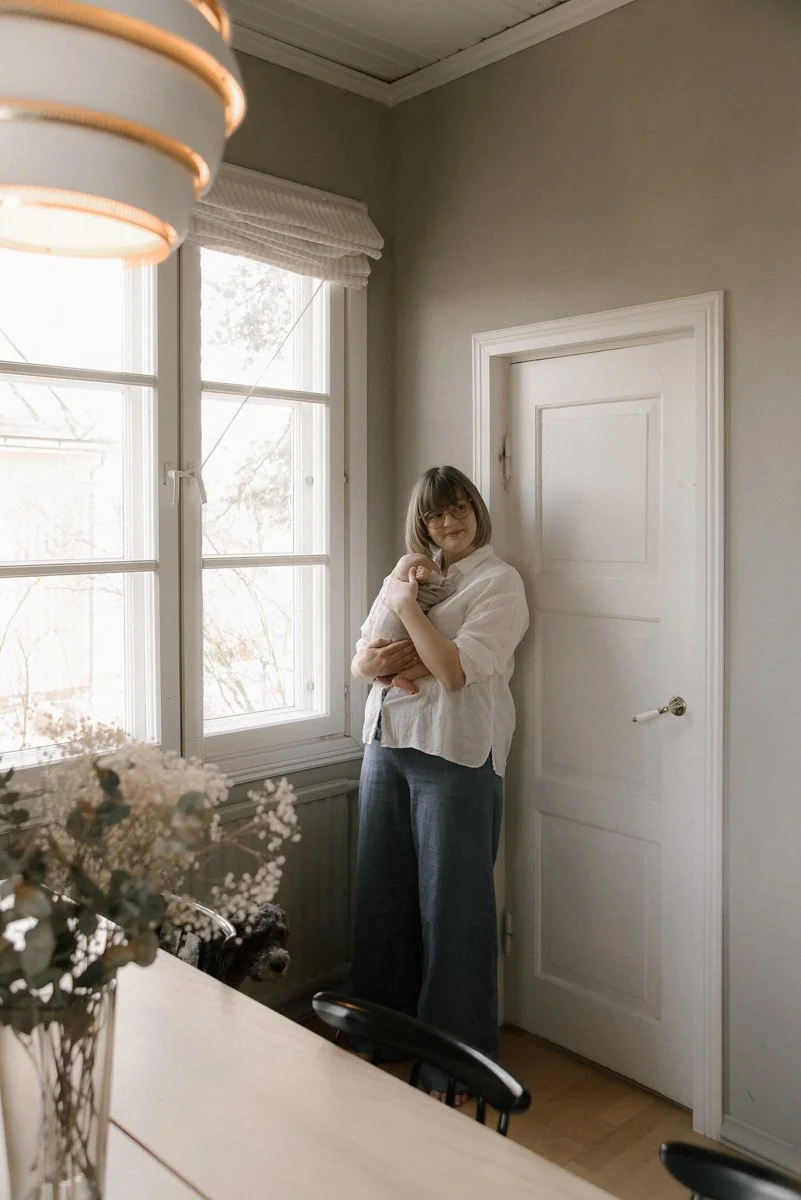 Nainen seisoo ikkunan vieressä keittiössä, pitelee vauvaa sylissään. Taustalla valkoinen ovi ja ikkunat, keittiö on valoisa ja vaaleasävyinen. Vauvakuvaus kotona.