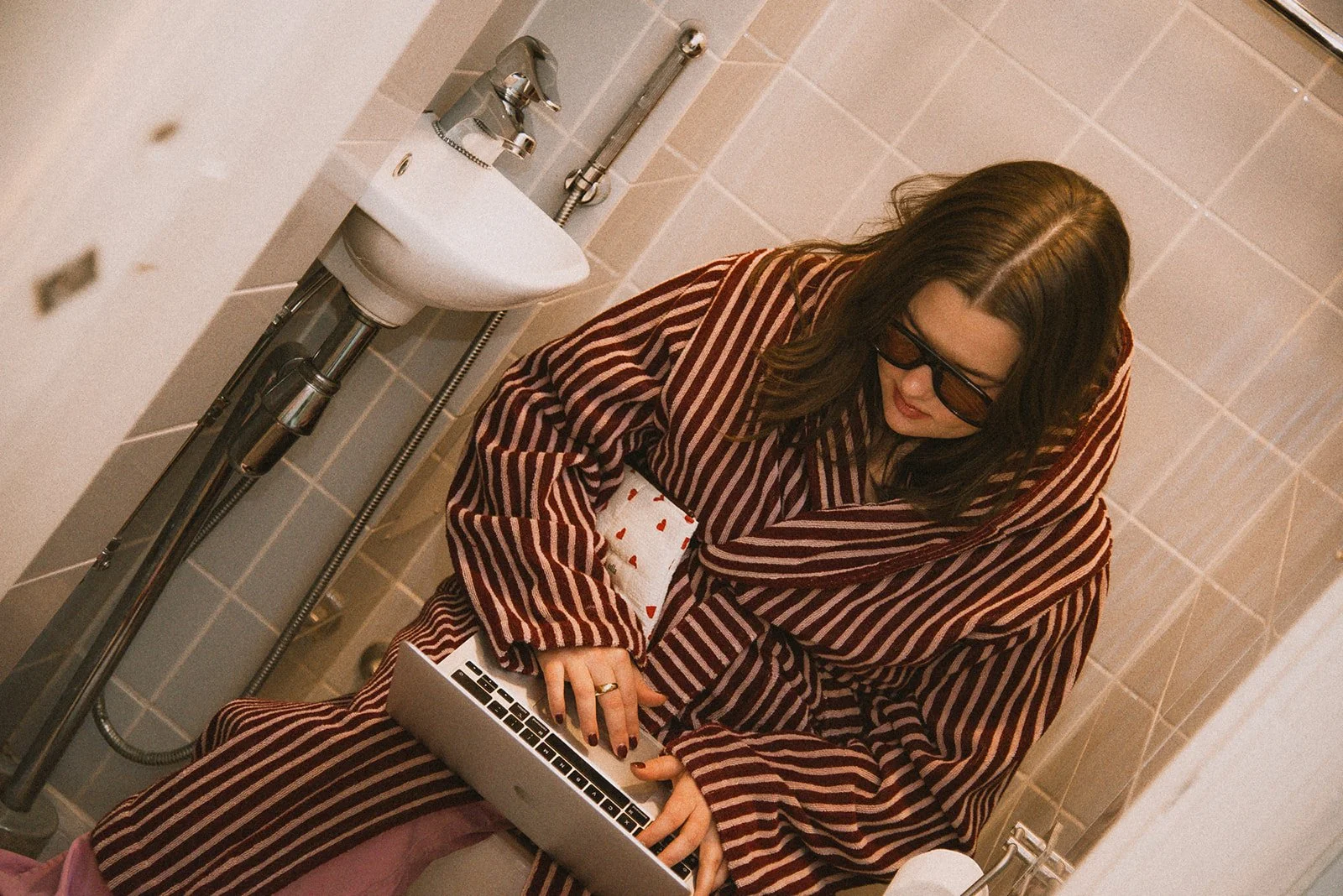 Nainen näpyttelee tietokonetta kylpyhuoneessa kylpytakki päällään. Kuvannut brändikuvaaja Heidi Väisänen.